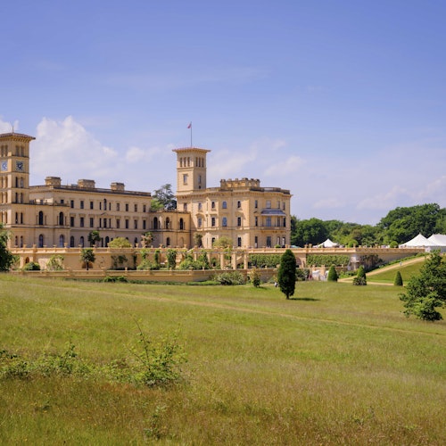 Prostrana, povijesna zgrada s tornjevima, okružena zelenim travnjakom i drvećem pod čistim plavim nebom.