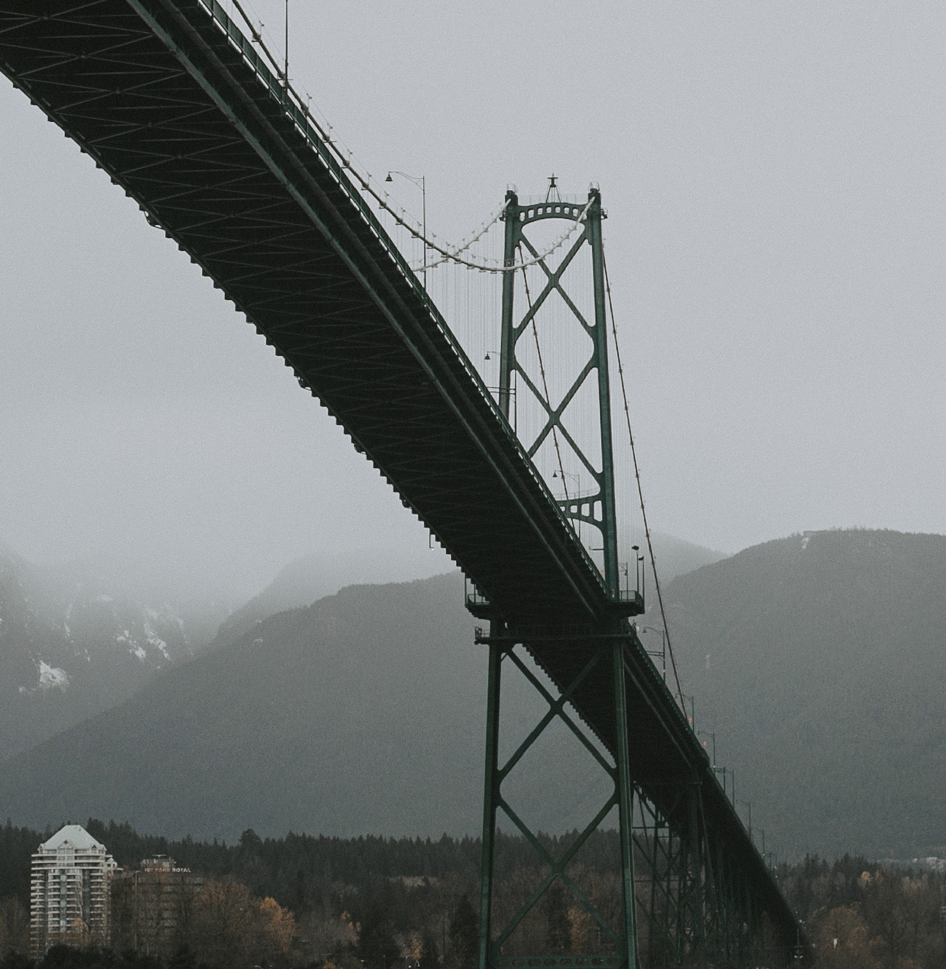 Lions Gate Bridge in Vancouver, Canada, seen from below on a foggy day, with its green steel structure stretching across misty mountains.