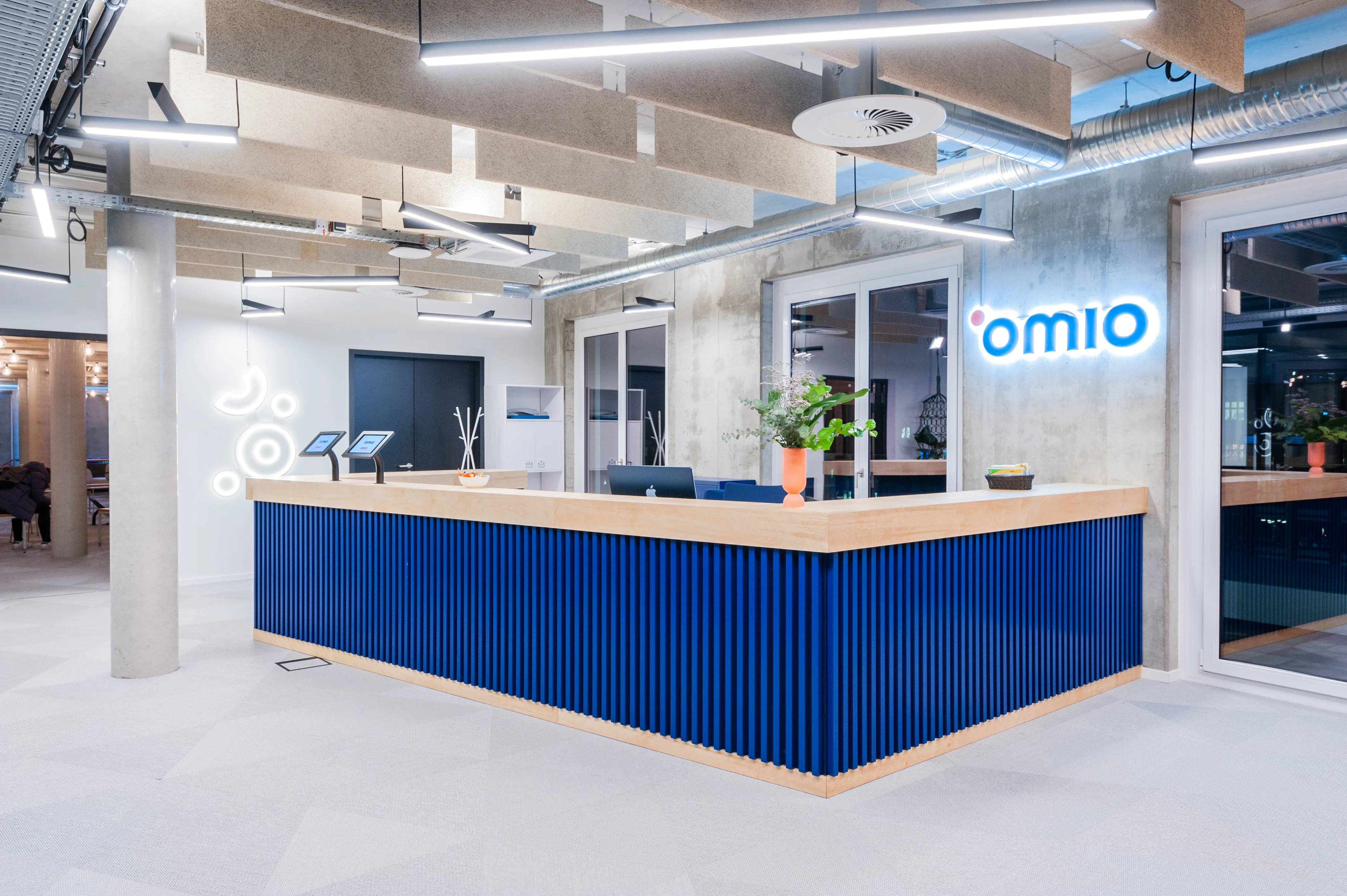 Reception desk at OMIO clad in royal-blue vertical slats with a wooden top, an illuminated white Omio logo on a concrete wall and wood-grain acoustic baffles overhead.