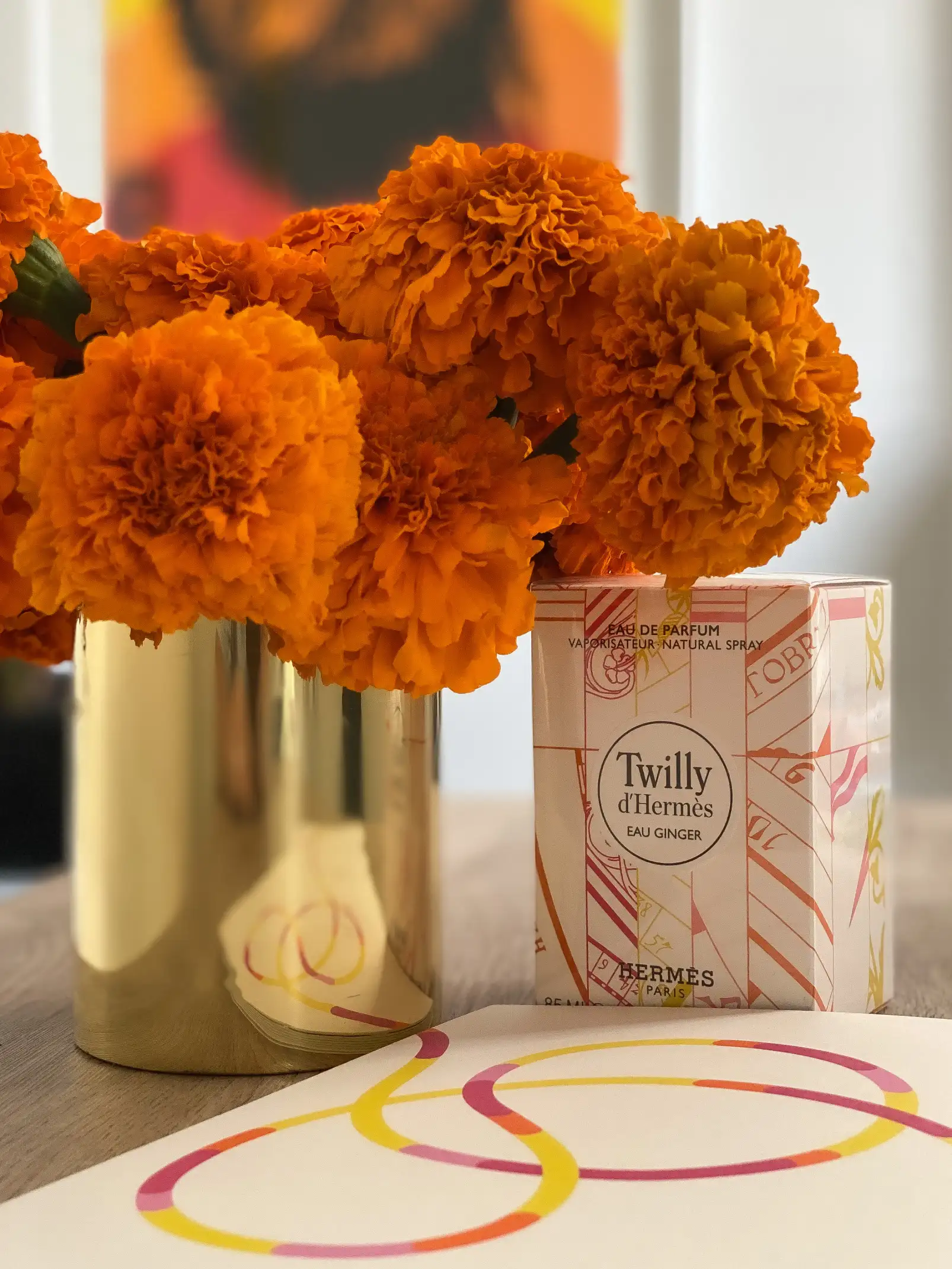 Ramo de flores de cempasúchil de color naranja intenso en un jarrón dorado junto a una caja del perfume Twilly d’Hermès Eau Ginger. La escena se completa con una hoja decorativa con líneas curvas en tonos rosa, amarillo y naranja, creando una composición cálida y elegante sobre una mesa de madera clara.