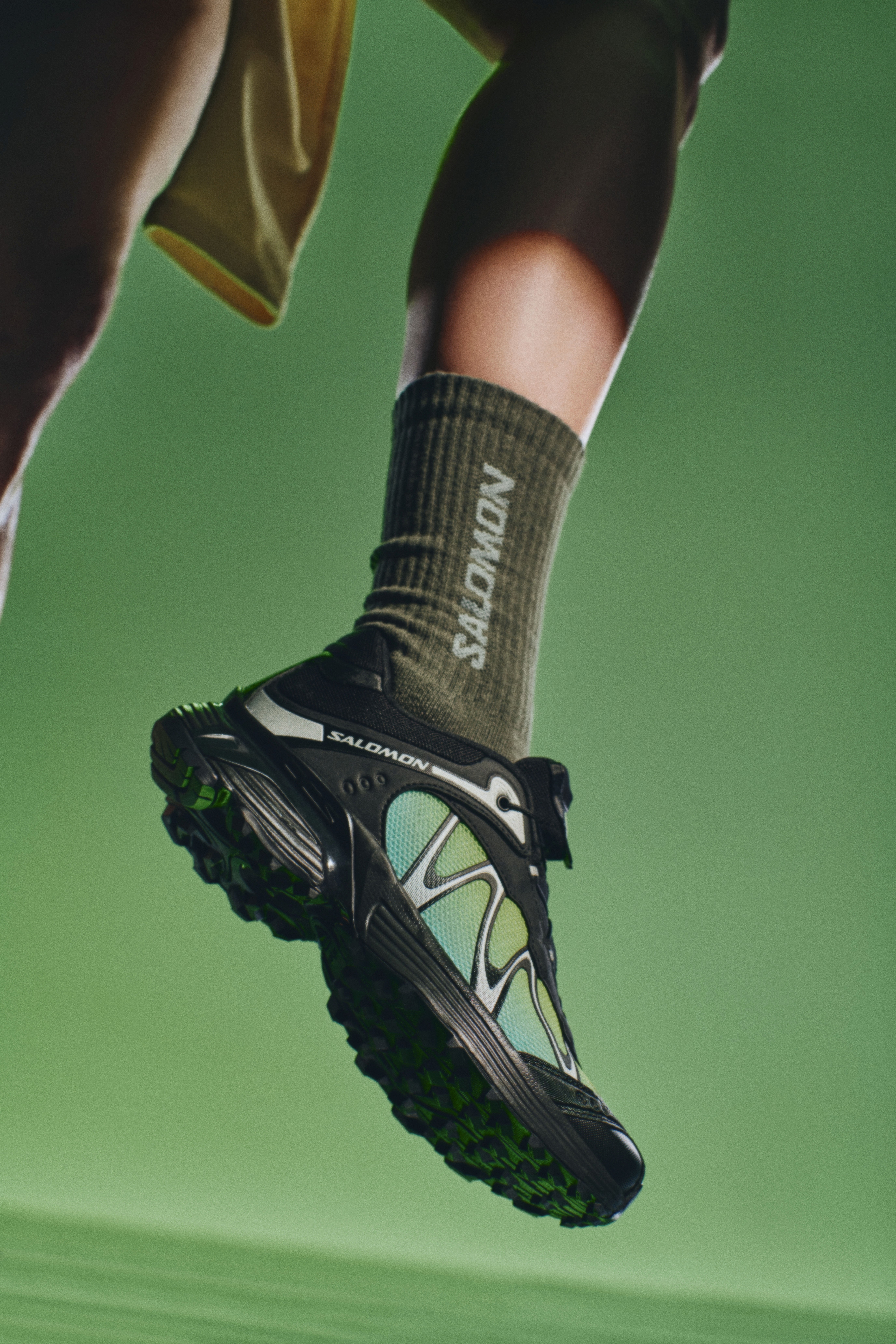 Un pied sur fond vert portant une basket Salomon noire et verte