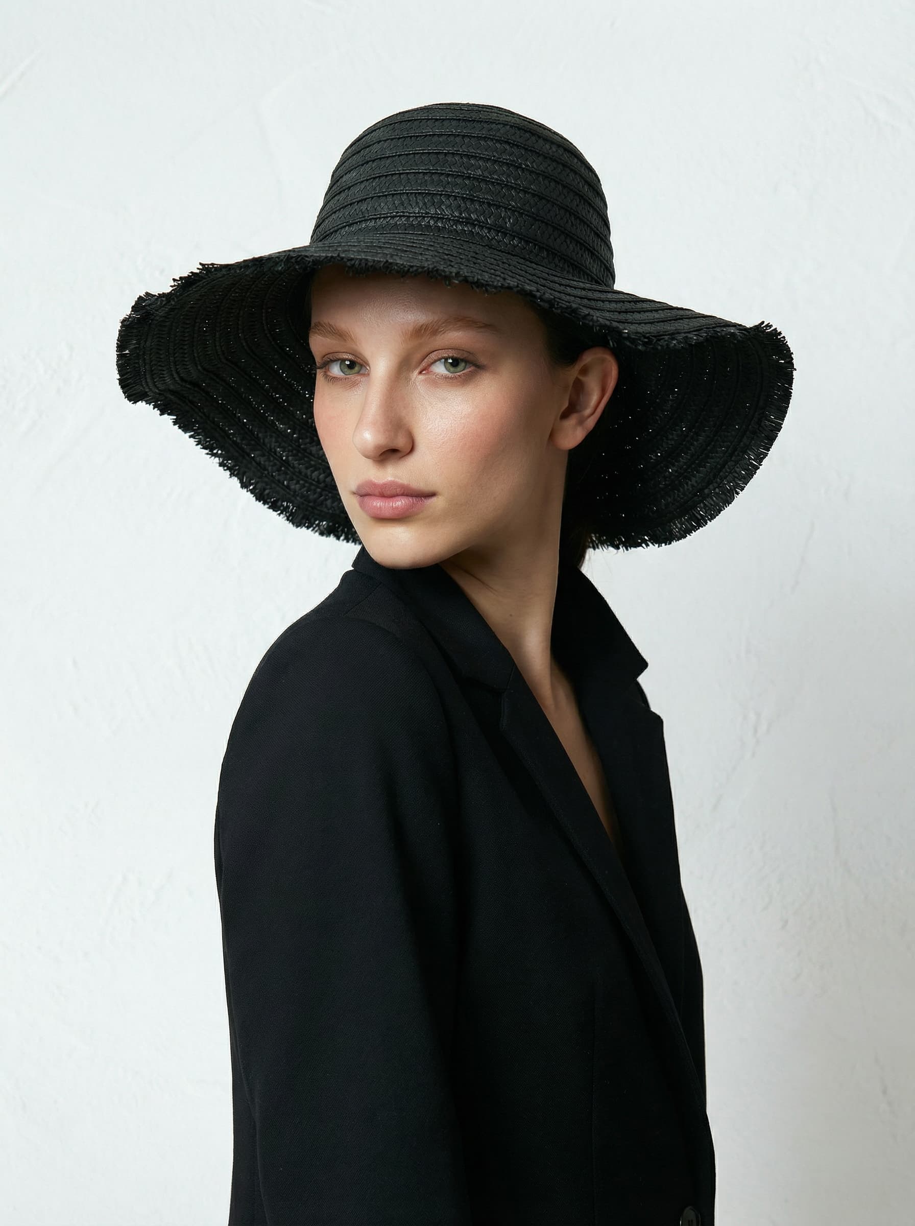 A person wearing a wide-brimmed black sun hat and a black blazer poses against a plain white background.