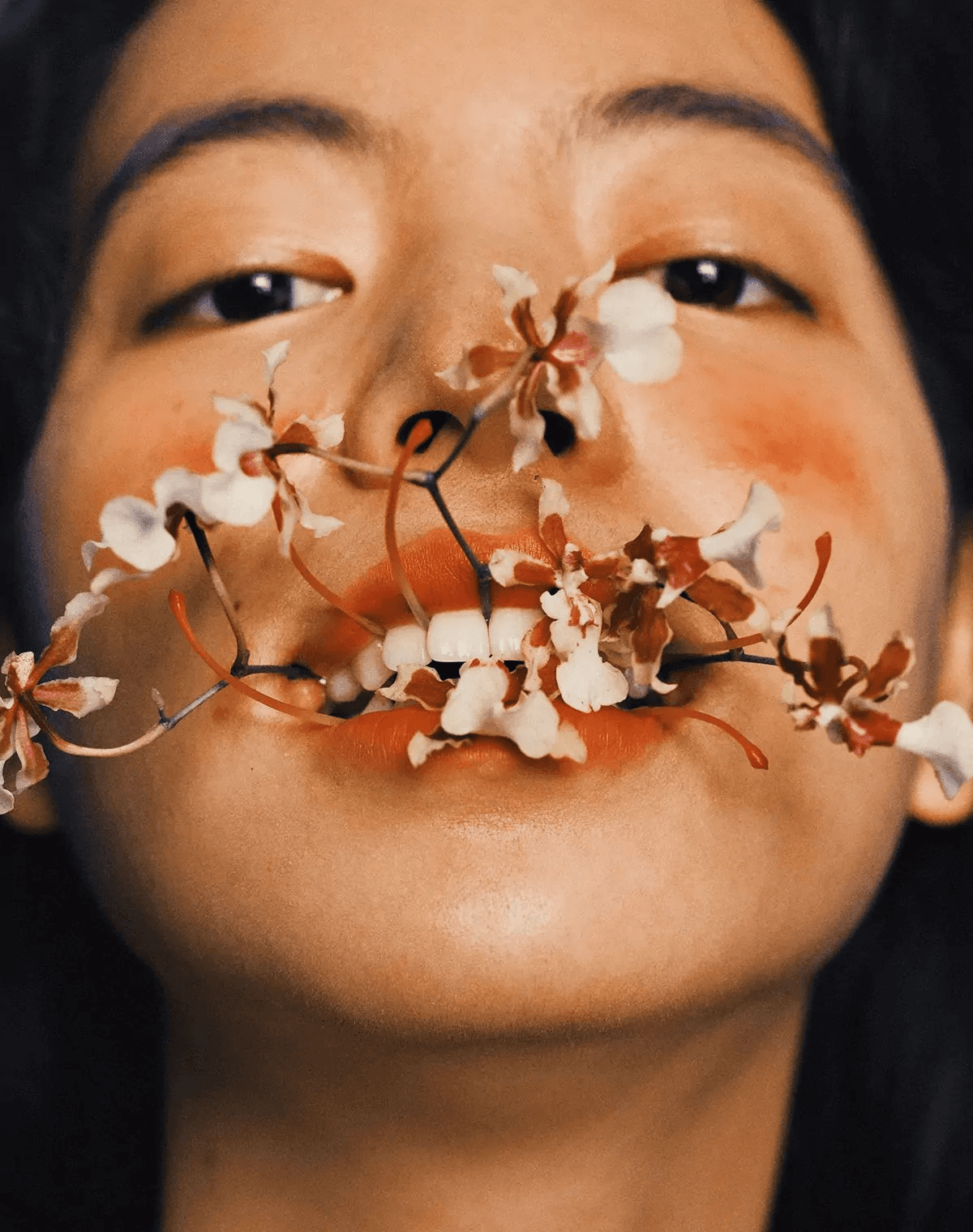 A black-haired woman looking at the camera while biting a flower.
