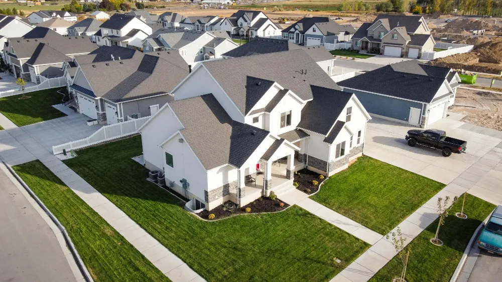 Angled aerial view of a modern suburban home with a manicured lawn, driveway, and surrounding neighborhood visible in the background. This perspective reflects real estate drone videography, allowing a drone videographer to showcase the home’s curb appeal and its position within the community.