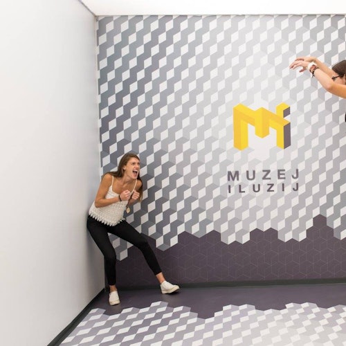 A woman stands against a patterned wall, appearing scared as a disembodied hand reaches down. The wall reads "Muzej Iluzij."
