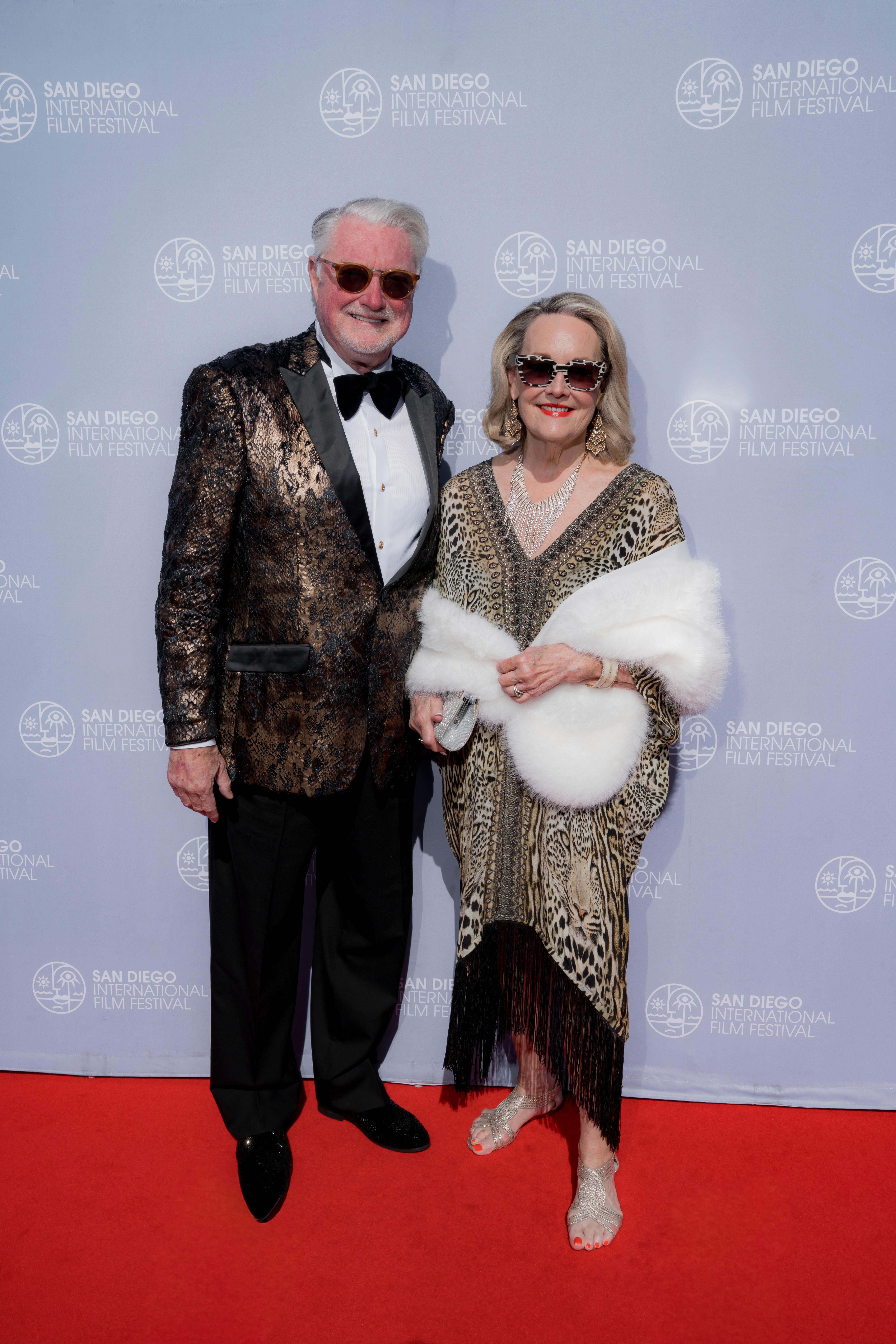 A couple posing together, the man in a metallic patterned blazer and the woman in a leopard-print dress with a white fur wrap.