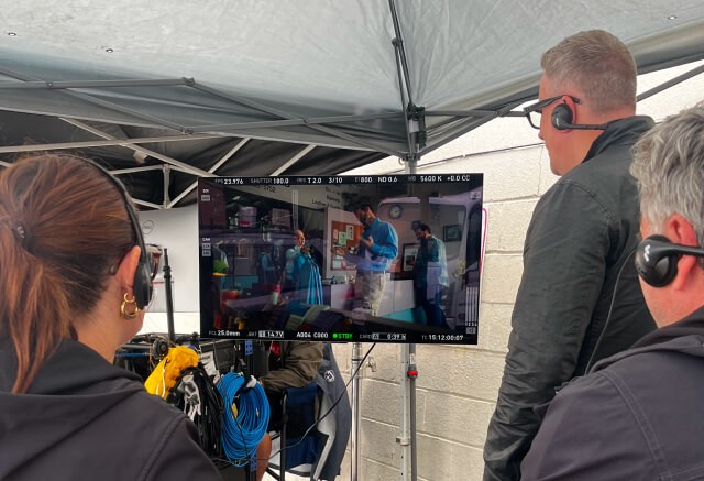 Three people watch a monitor showing a hospital scene being filmed, with equipment and cables visible under a canopy on set—a behind-the-scenes look at creative branding and storytelling in action.