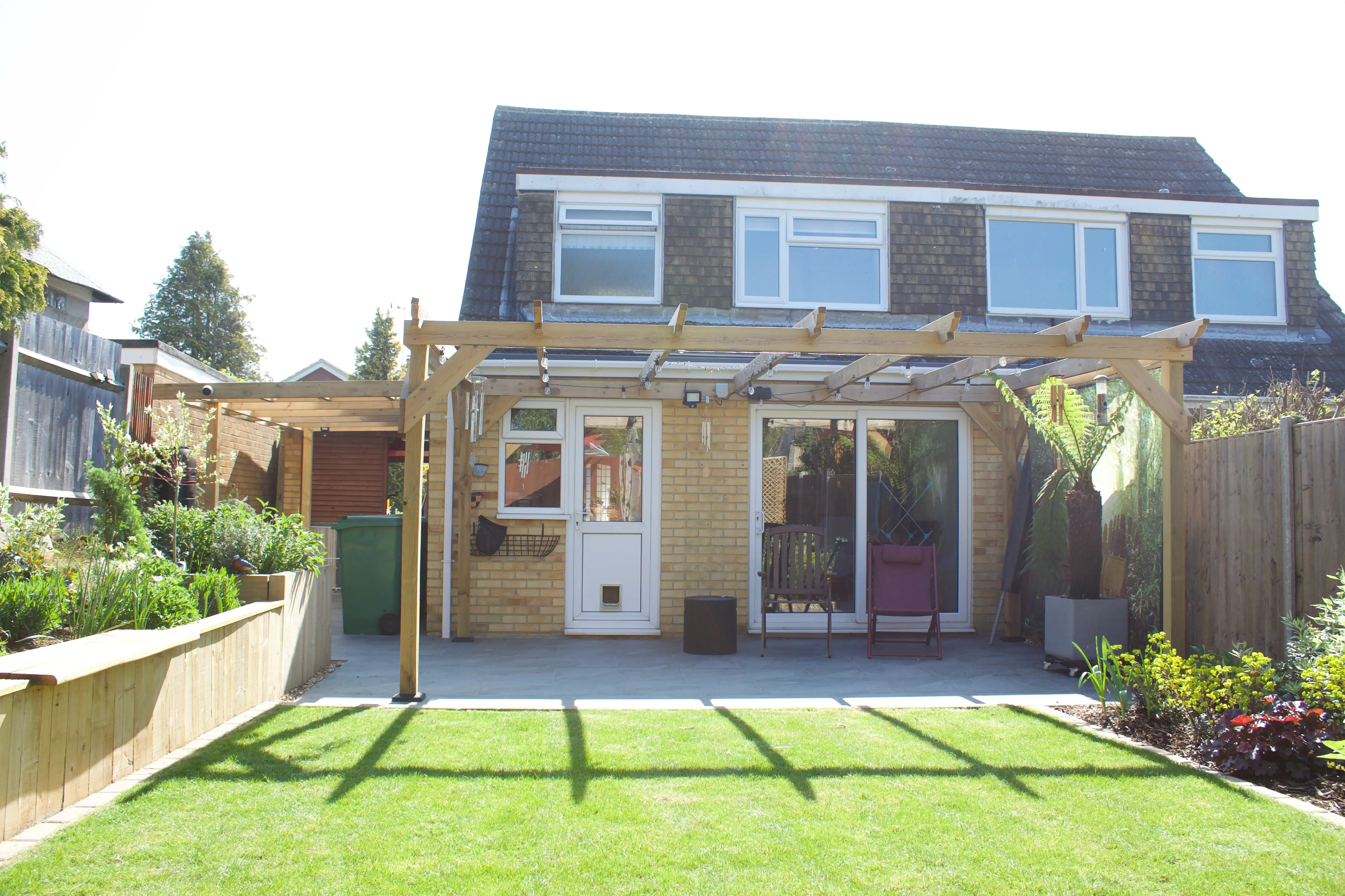A two-story house with a garden, featuring a patio and greenery in the foreground under a clear blue sky.