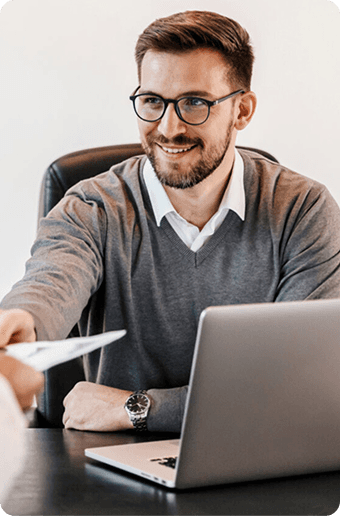 A man in a sweater smiles while handing over a document from a desk with a laptop.