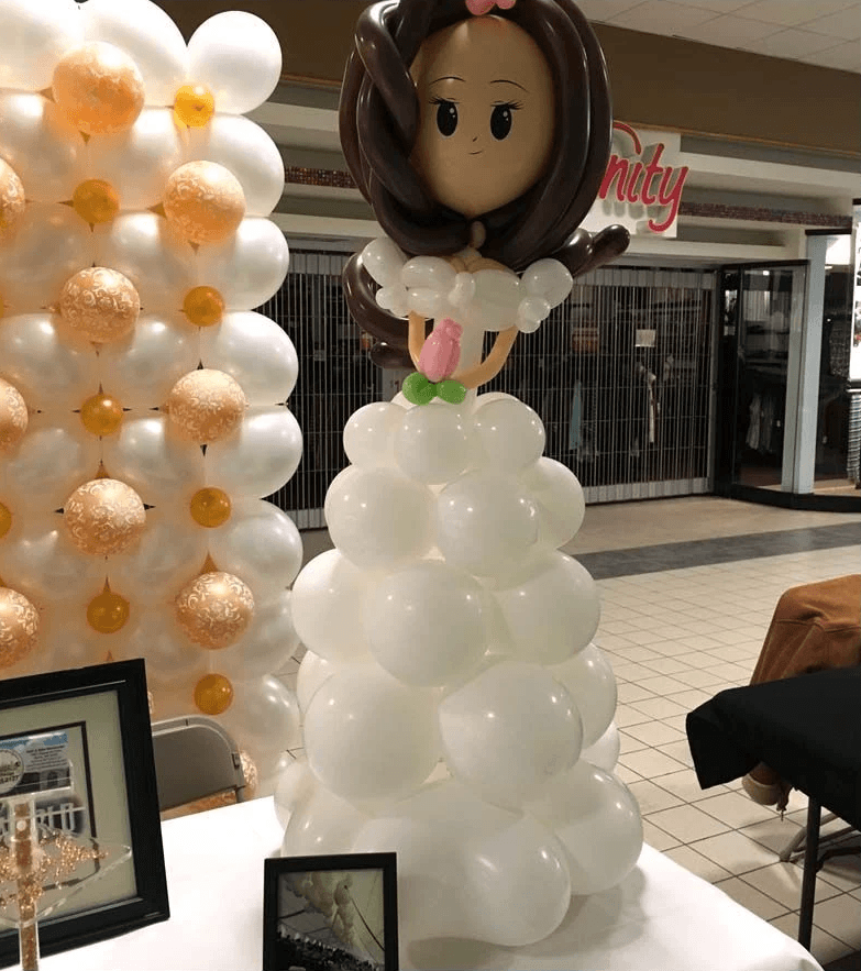 A large balloon sculpture of a bride with a white gown and brown hair is displayed in a shopping mall, with art frames placed on a table in the foreground.