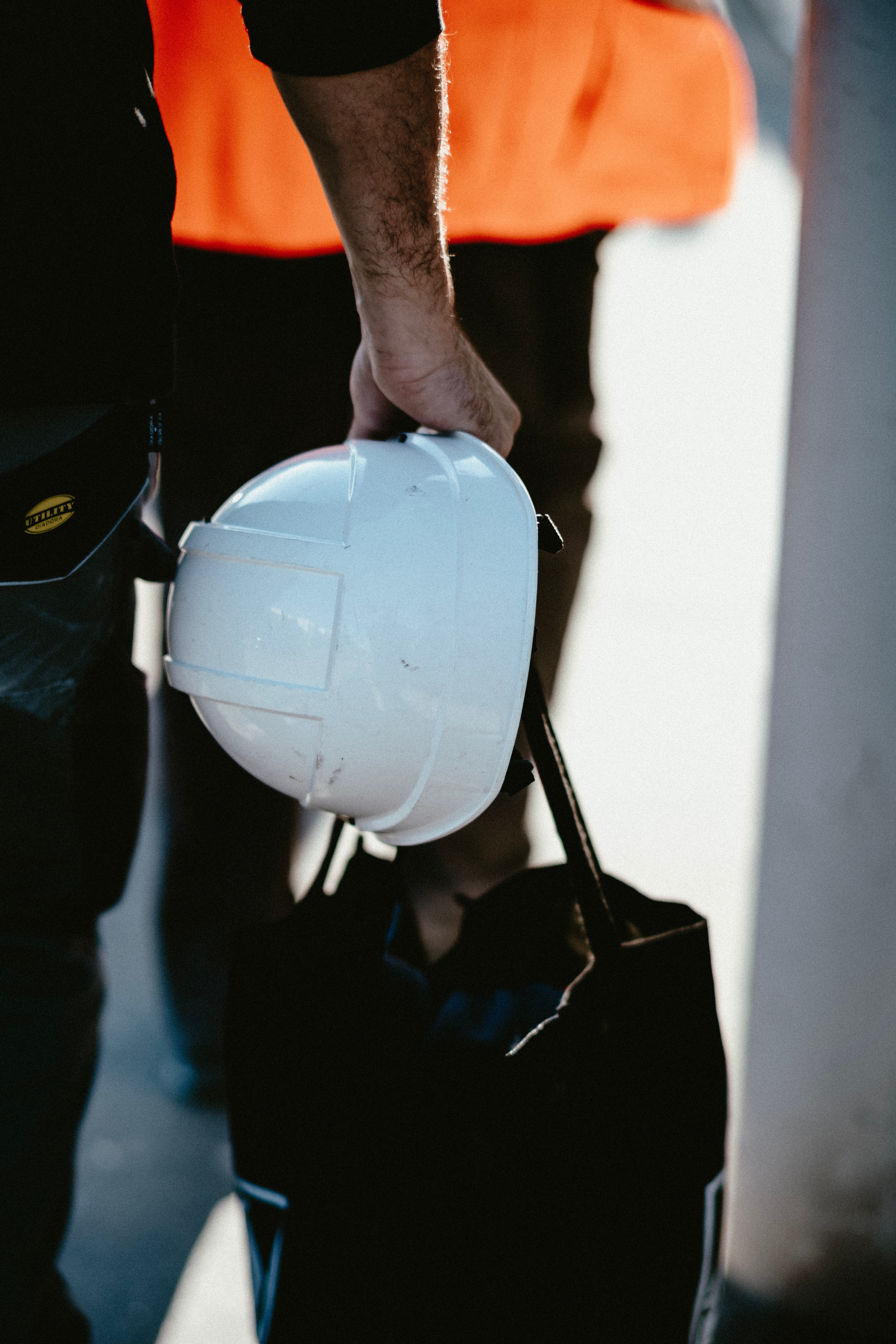 A close up of a white hard hat and black work back held in the hand of a person in an orange vest