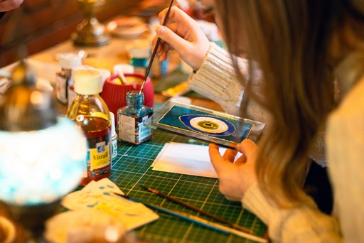 A person crafting a Turkish mosaic lamp, carefully applying multicolored beads to the ornament. The background shows a workshop setting with tools hanging on the wall.