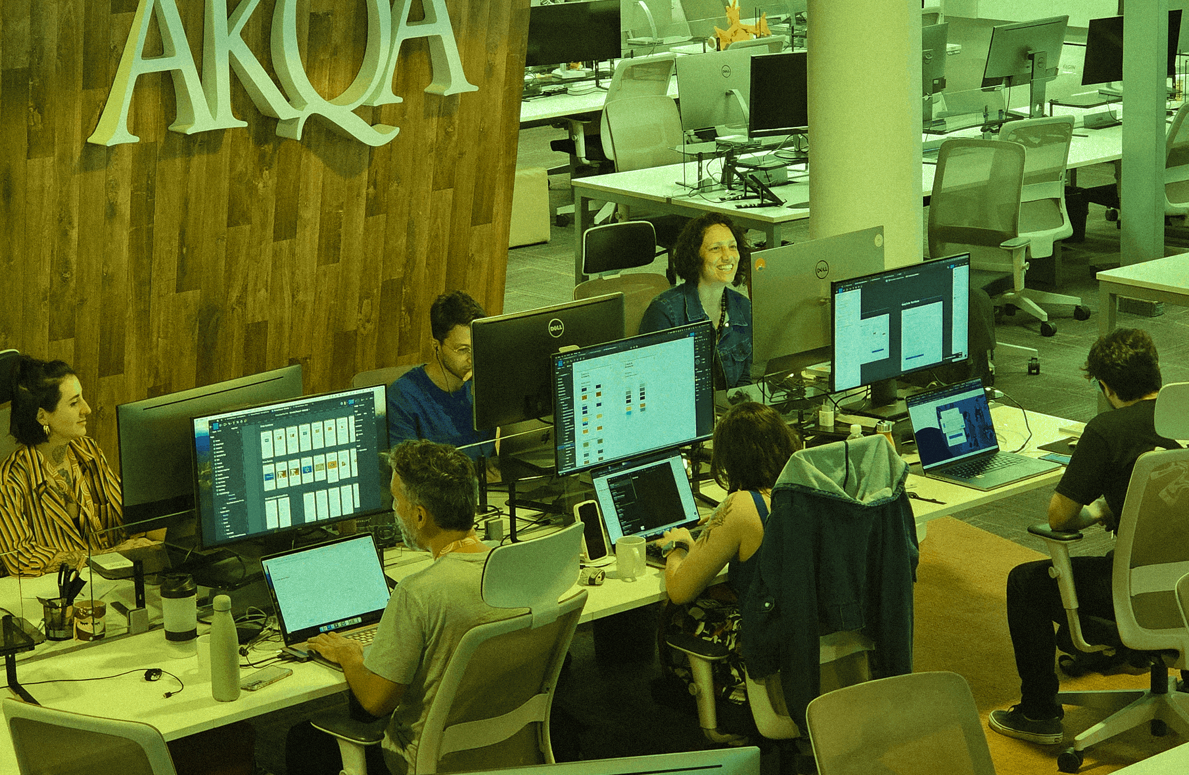 Design professionals working at their computer stations in an open-plan office, with a large wooden AKQA sign on the wall behind them.