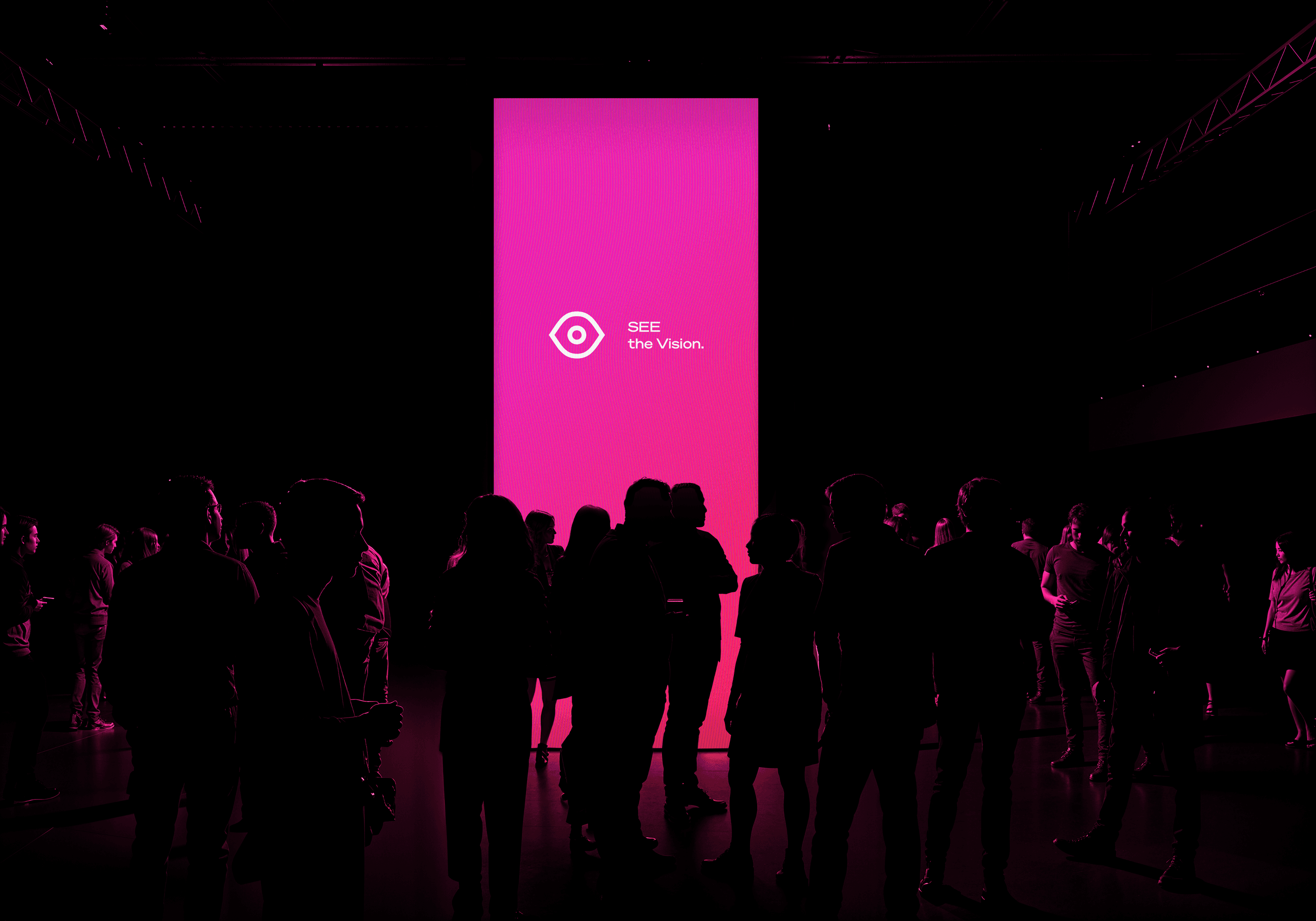 Tall magenta vertical LED banner with "SEE the Vision" typography glowing in a dark event hall surrounded by attendees.