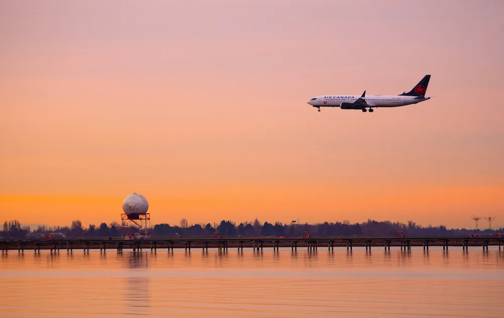 A plane is flying above a body of water. There is a sunset on the background.