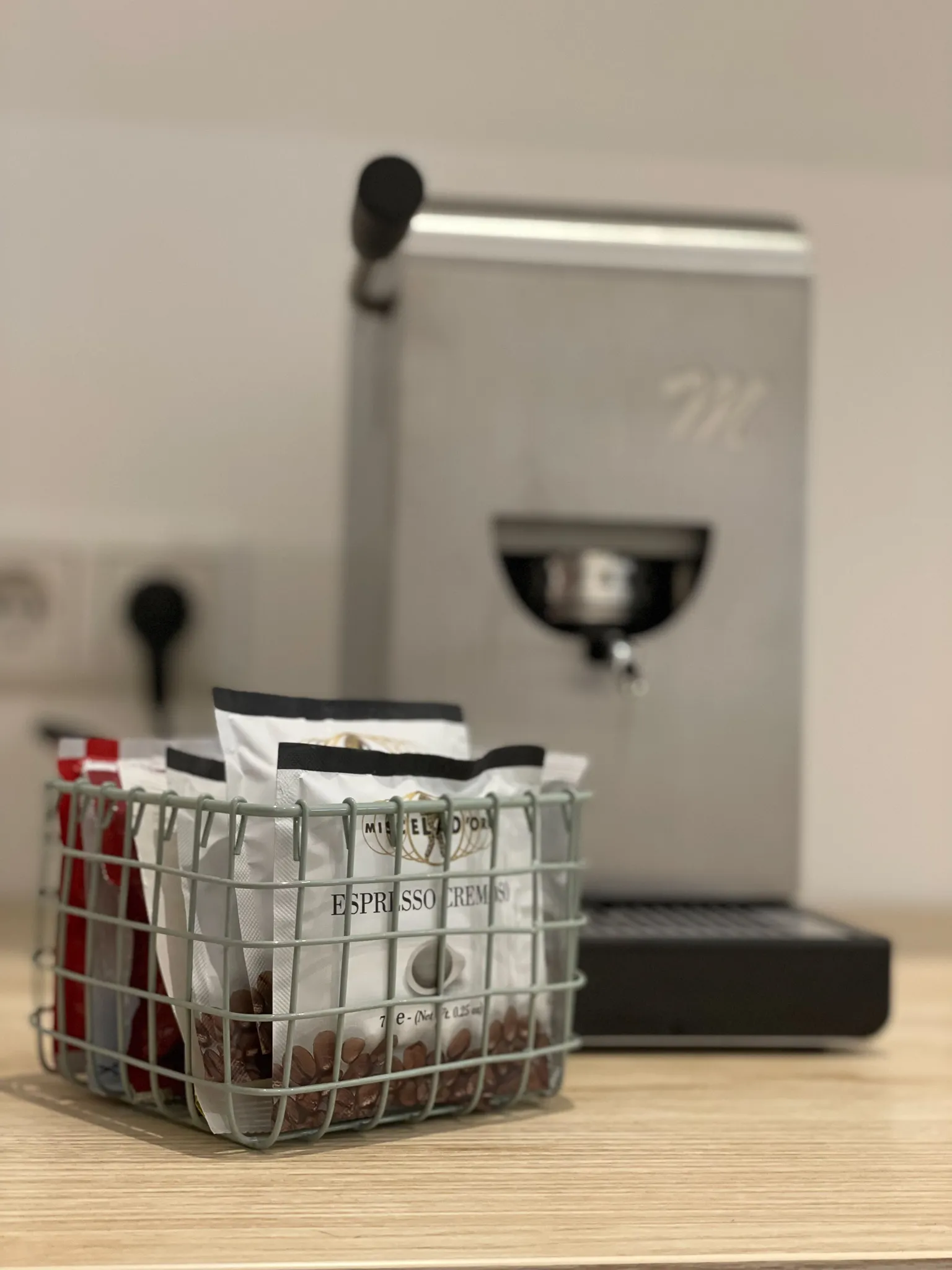 „Metallene Kaffeemaschine auf einer Holztheke mit einem Korb voller Espressobohnen-Päckchen im Vordergrund; minimalistisches und gemütliches Küchenambiente.