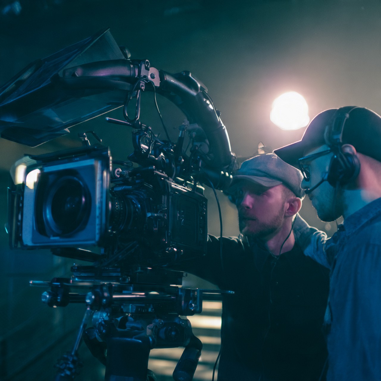 Two camera operators in a dimly lit studio adjust a large video camera. They wear headsets and caps, focusing intently. Mood is professional and focused.