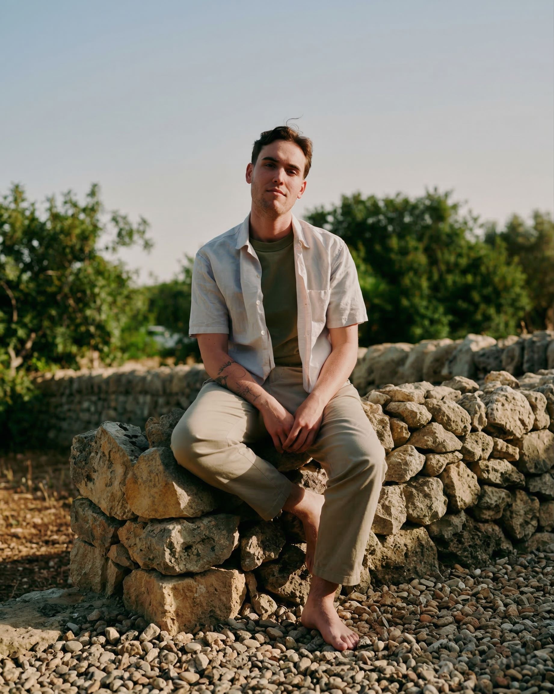 Marc sits on a stone wall with greenery in the background, wearing a light shirt and casual pants.