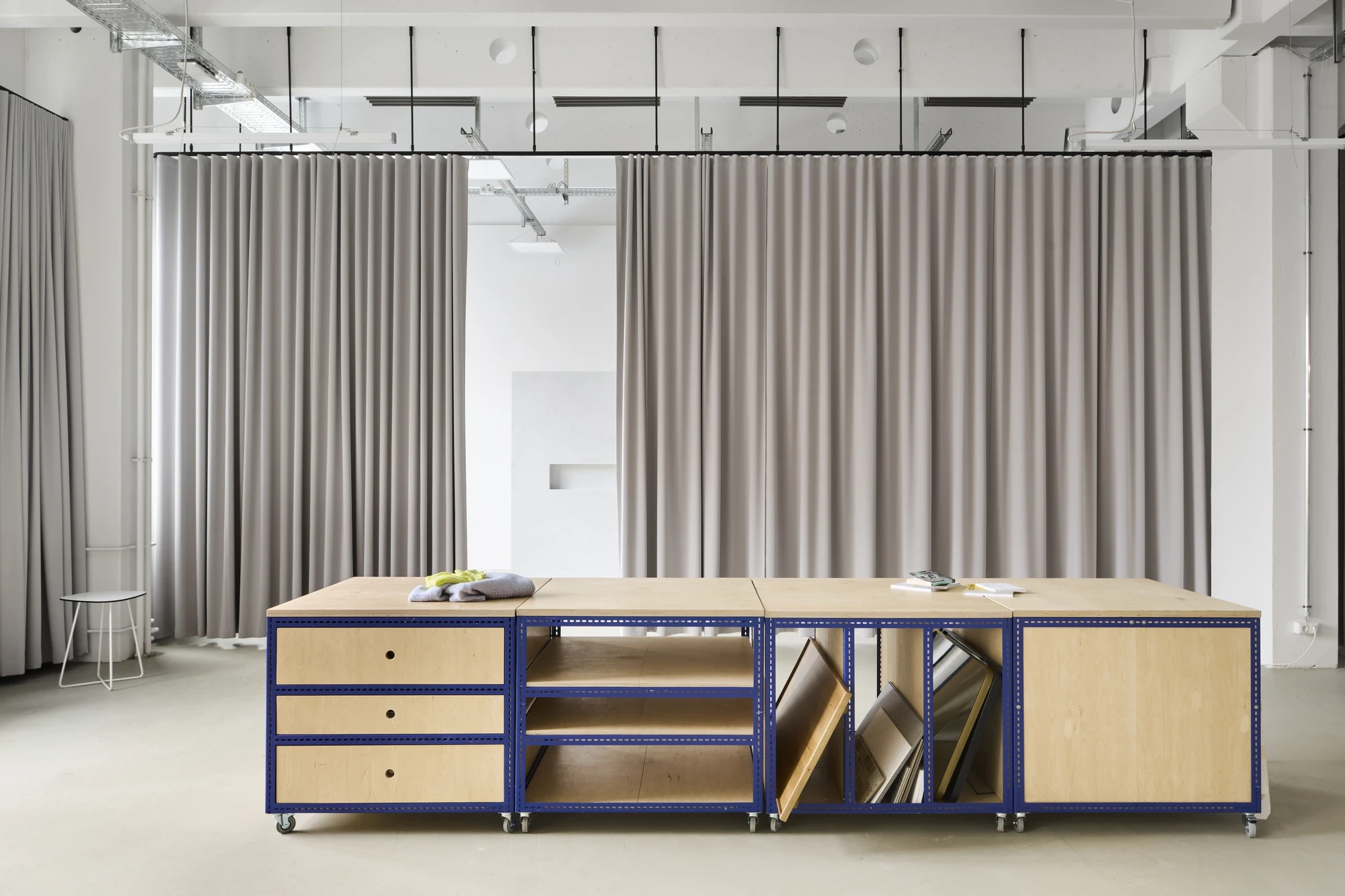 Modular wooden workbench with cobalt-blue metal frame, drawers and storage compartments, set against a backdrop of beige drapes.