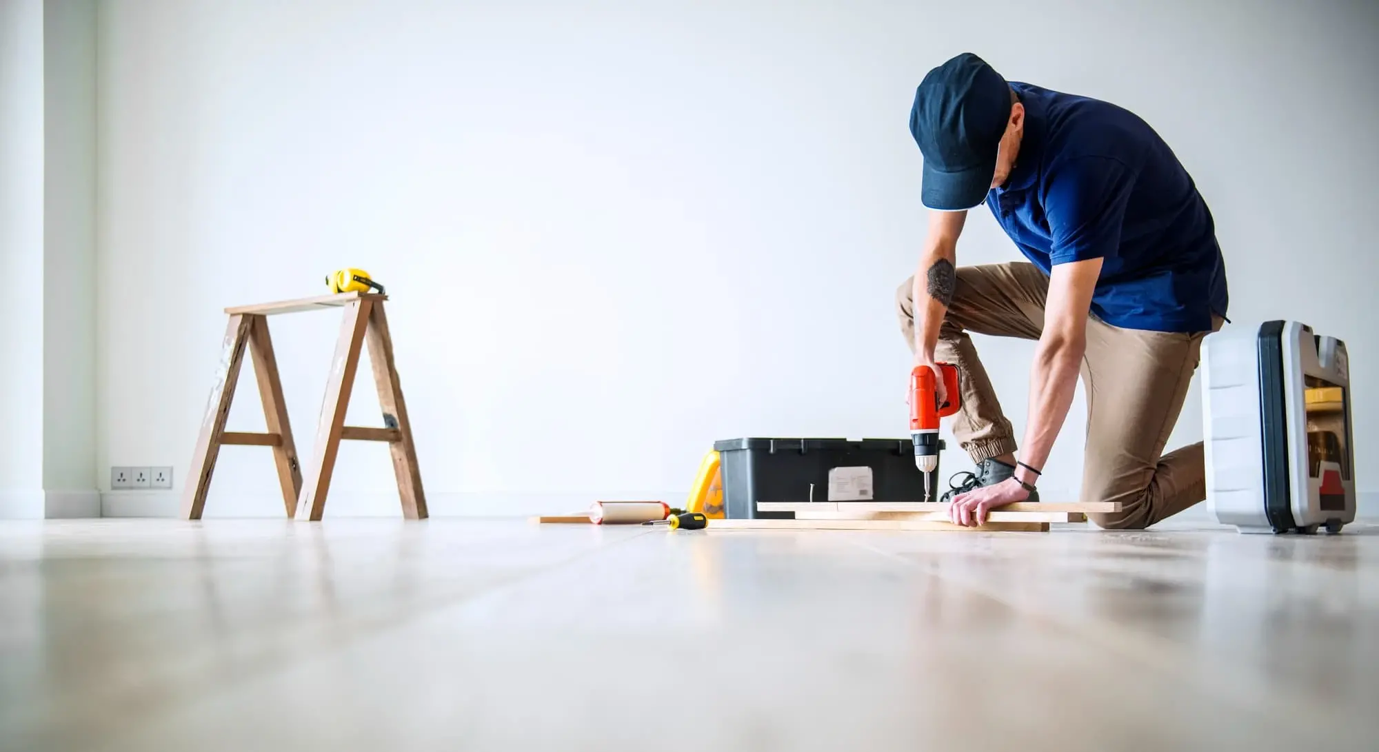 A man kneels on a hardwood floor using a power drill to make home improvements, surrounded by tools and a ladder. The image represents using cash-out refinancing from Chris Lewis Home Loans to fund renovations and add value to your home.