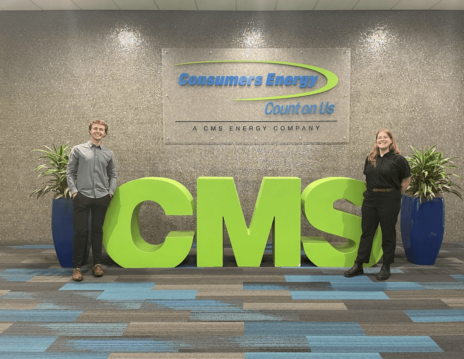 Two individuals stand beside large green "CMS" letters in a modern office space, with plants and a wall sign in the background.