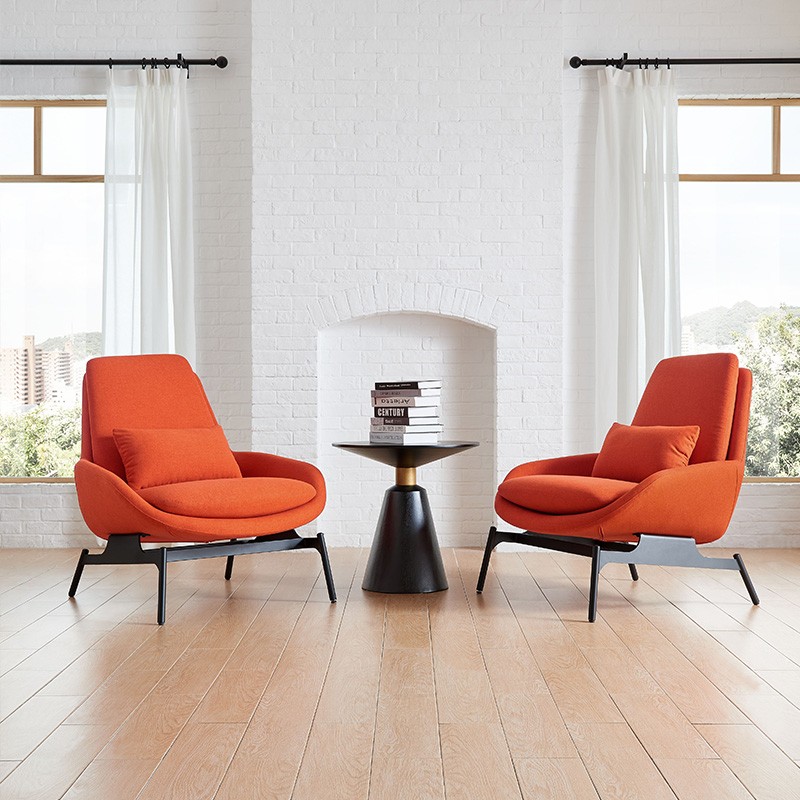 Symmetrical living room arrangement featuring two orange mid-century modern lounge chairs with lumbar pillows, flanking a black conical pedestal side table in front of a white brick fireplace and large windows.