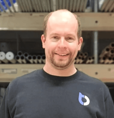 Blid mann med skjegg i blå genser med BlueTech logo foran hyller med rørdeler