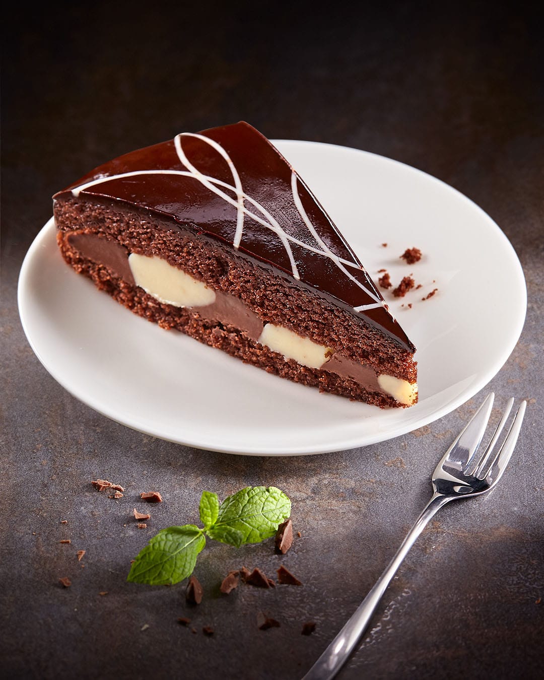 Kawałek eleganckiego ciasta czekoladowego z błyszczącą polewą ganache, zdobiony skomplikowanymi białymi wzorami z lukru i kremowym nadzieniem, podawany na białym talerzu obok gałązki świeżej mięty i wiórków czekoladowych, z widelcem położonym w pobliżu.