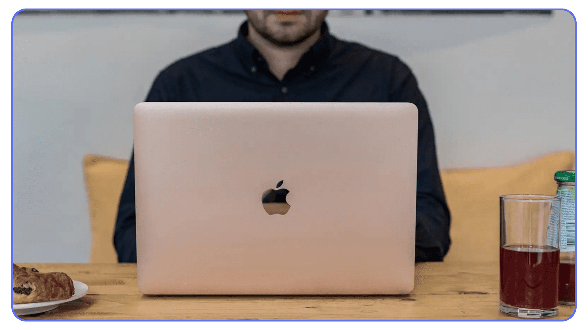 Person working on a rose-gold MacBook at a table with drinks and snacks nearby.