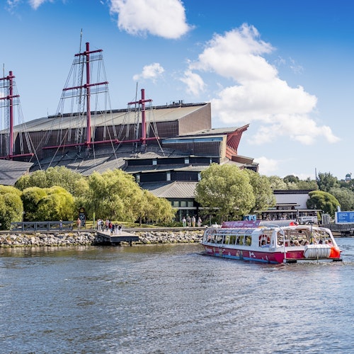 A red and white tour boat on a river, with a large building and lush trees along the riverbank under a blue, partly cloudy sky.