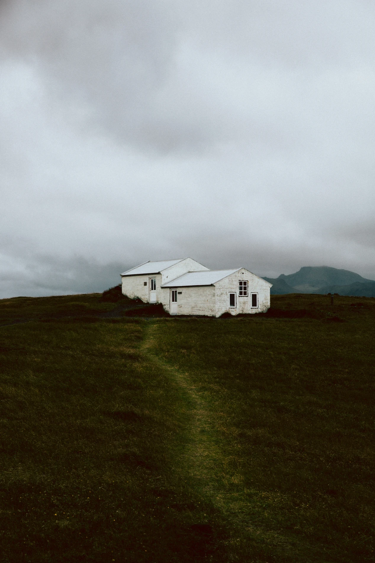 A small white farmhouse alone on a green slope under heavy cloud