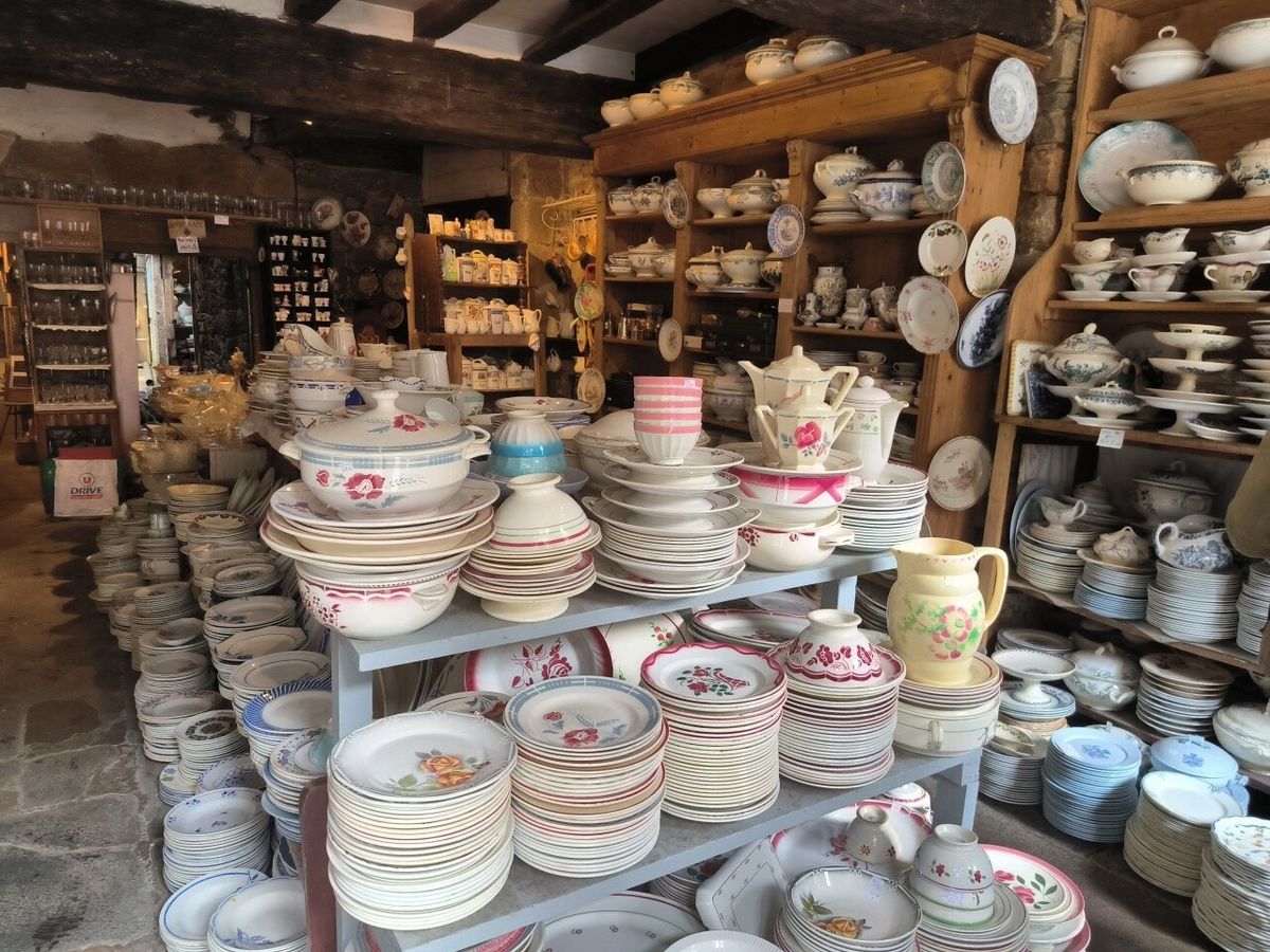 Intérieur d'une brocante rustique avec poutres apparentes, étagères et présentoirs en bois chargés d'assiettes, soupières, théières et pichets en faïence ancienne aux motifs floraux roses et bleus