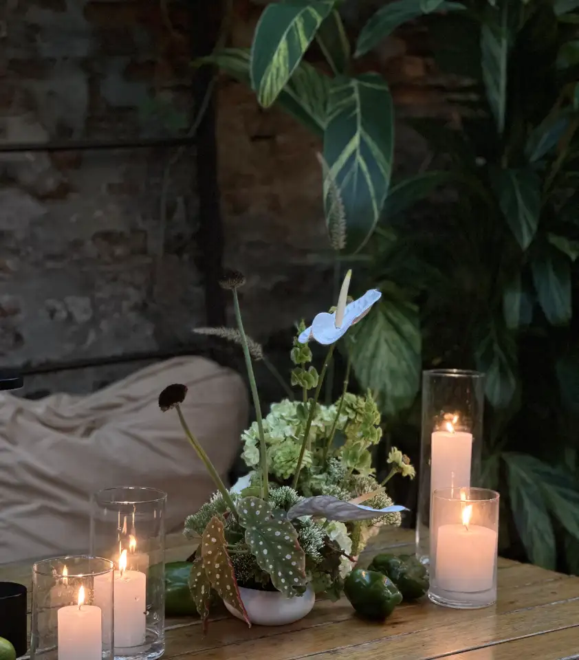 Centro de mesa con flores verdes y blancas, velas encendidas y vegetación tropical sobre mesa de madera para ambientación íntima y natural.