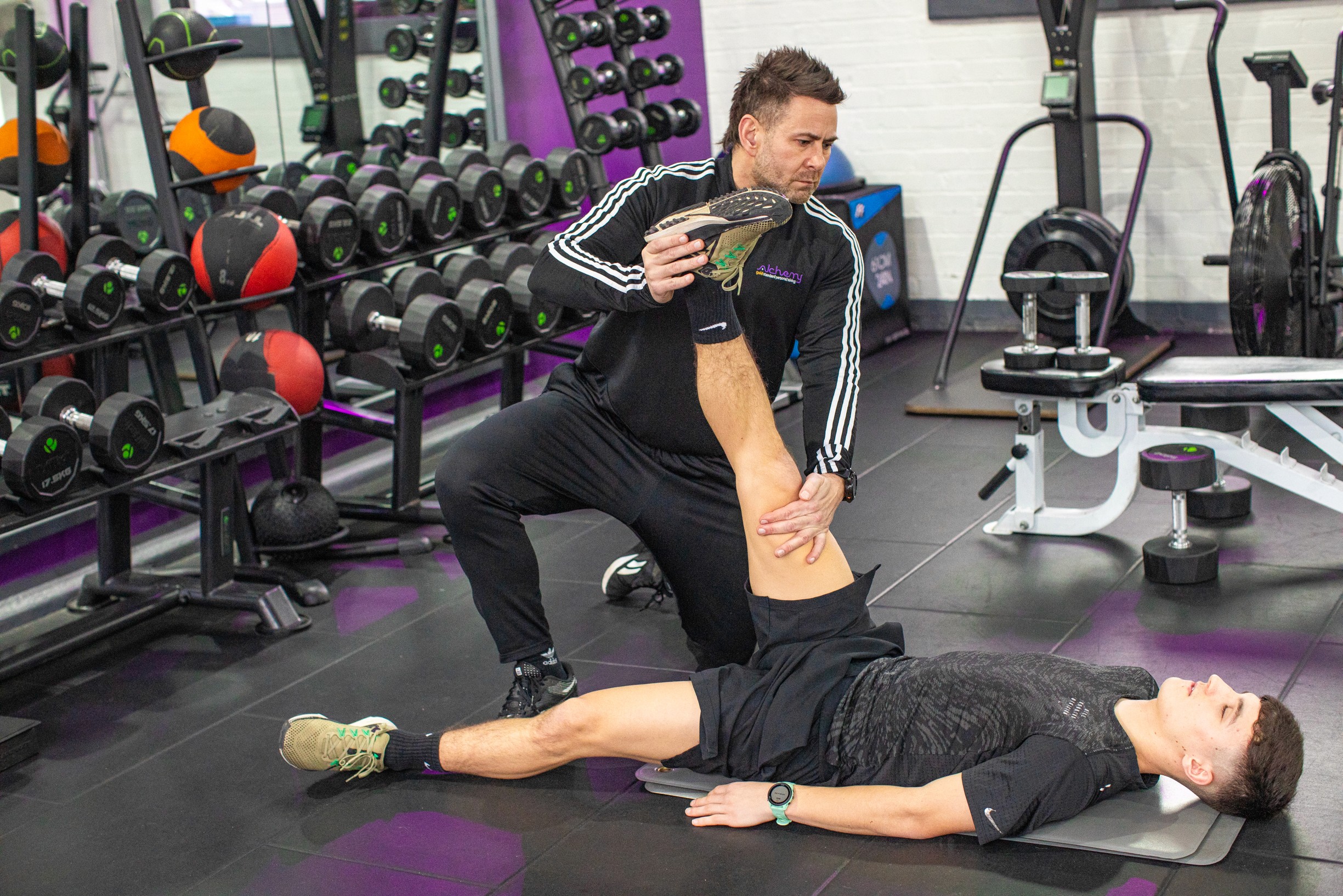 A personal trainer assists a young man with a leg mobility exercise in a well-equipped gym.