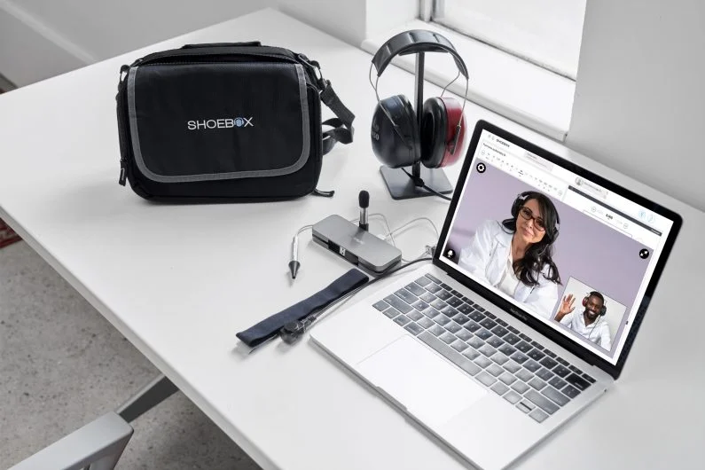 A laptop with a video call open, headphones, a camera, and a bag on a white desk.