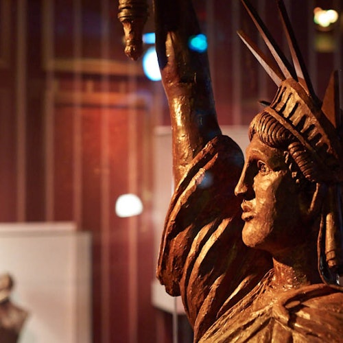 En närbild av en bronsstaty av Frihetsgudinnan i ett svagt upplyst utställningsutrymme med suddiga bakgrundselement.