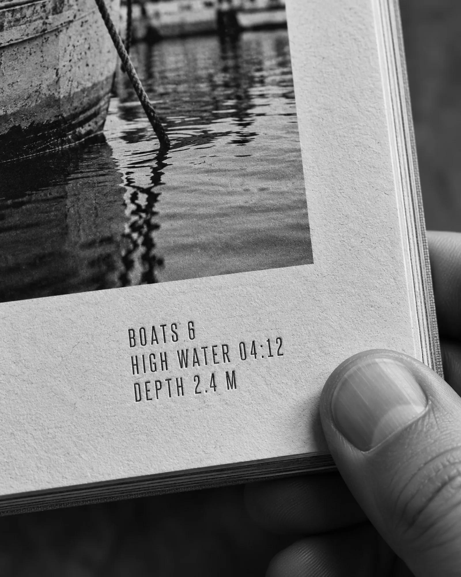 Close-up of a book spread corner showing a caption block in Valk Grotesk with boat count and high-water time, a thumb holding the page.