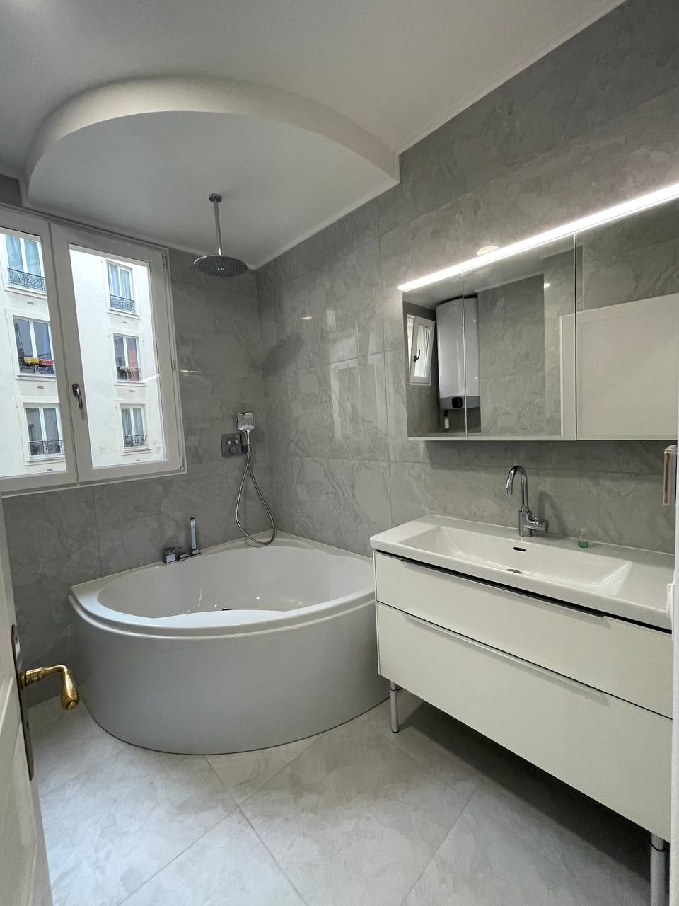 Salle de bain élégante avec baignoire d'angle, carrelage mural gris effet pierre et grand miroir éclairé.