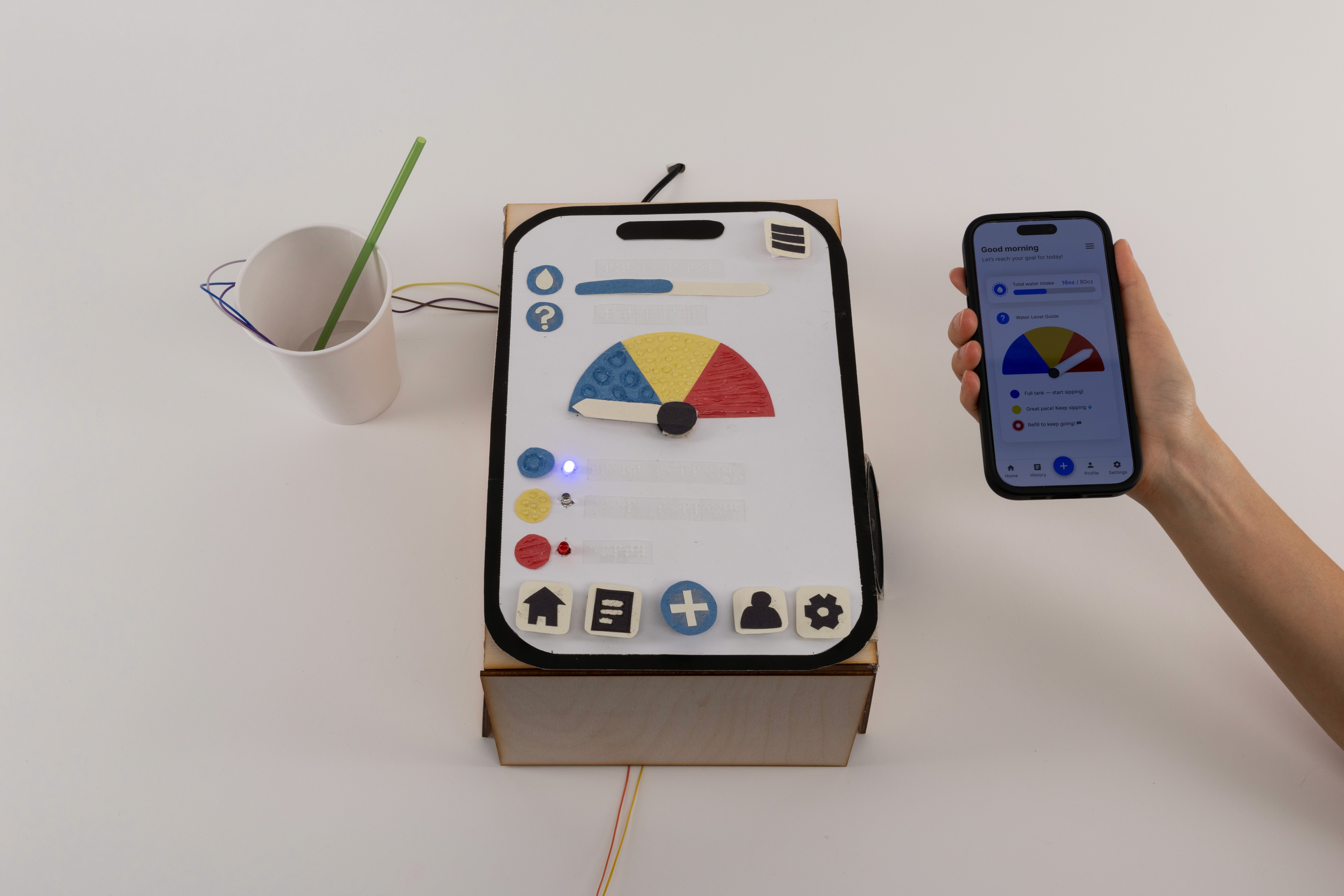 Sip 'n Sense product: a cup with a straw and wries. The wires run to a box shaped like a giant phone. Next the to the box a person's hand is holding a real phone. The image on the phone matches the look of the box.