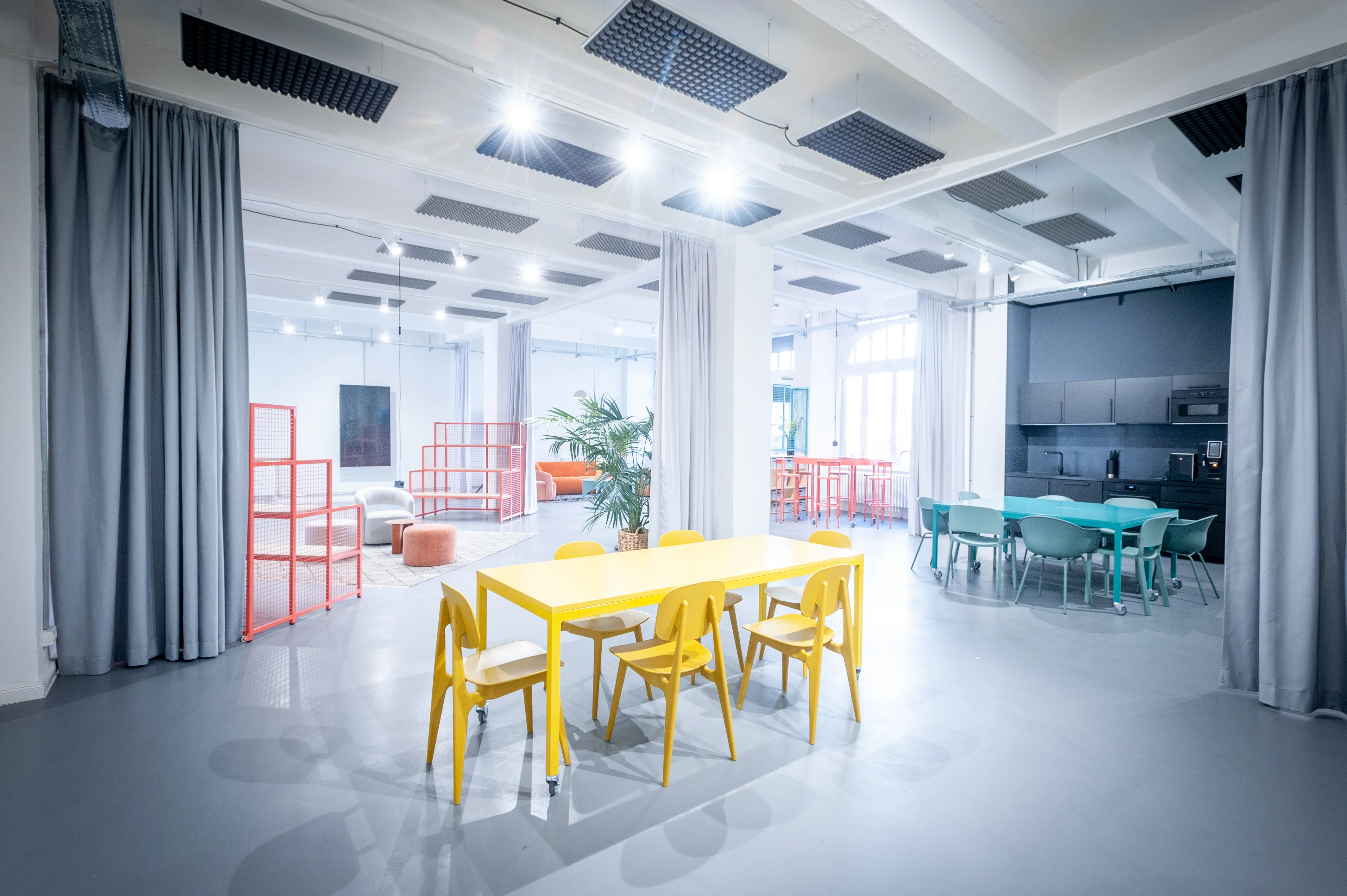 Overview of HEY CONTENT STUDIO with a yellow dining table group, turquoise meeting tables, coral wire shelves, grey curtain dividers and acoustic panels on a white ceiling.