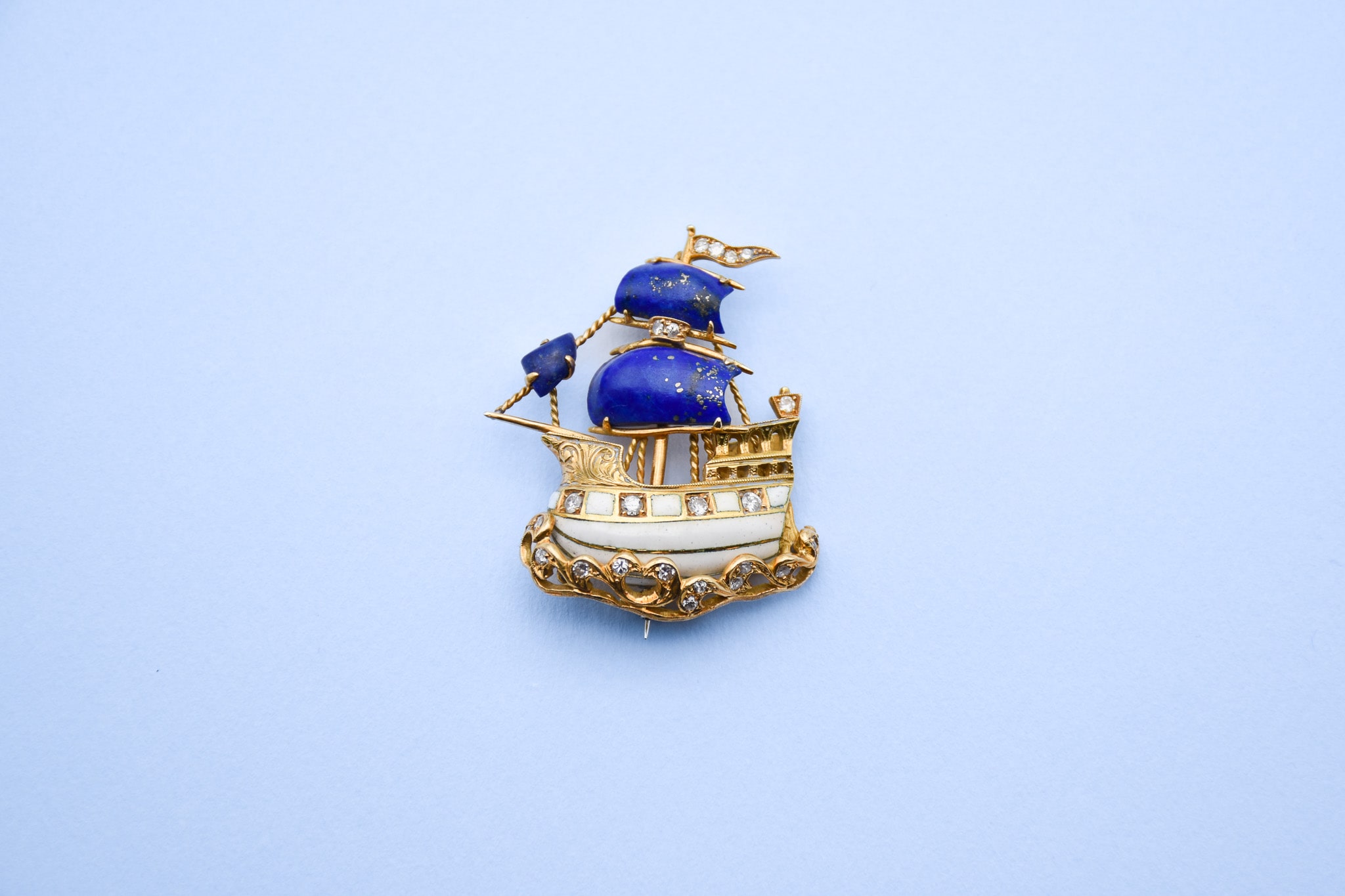 Vue de face d'une broche représentant un galion naviguant sur les eaux dont les voiles sont en lapis-lazuli et la coque en émail blanc ornée de petits diamants