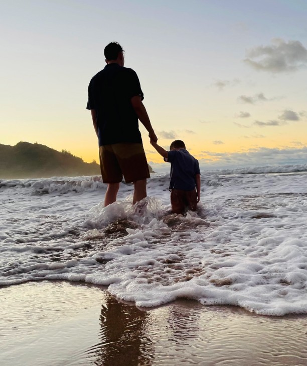 A serene beach scene at sunset featuring a person holding the hand of a young child as they stand together in the shallow surf. The ocean waves gently wash over their feet, with the horizon and distant hillside silhouetted against the pastel colors of the evening sky, creating peaceful and intimate moment by the sea.