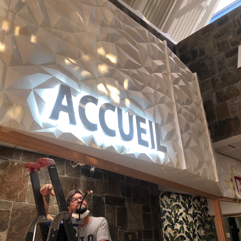 Un homme sur une échelle travaille sous une enseigne lumineuse sur laquelle on peut lire « ACCUEIL » en lettres grasses. Le mur est orné d'un motif géométrique blanc, créant un look moderne.