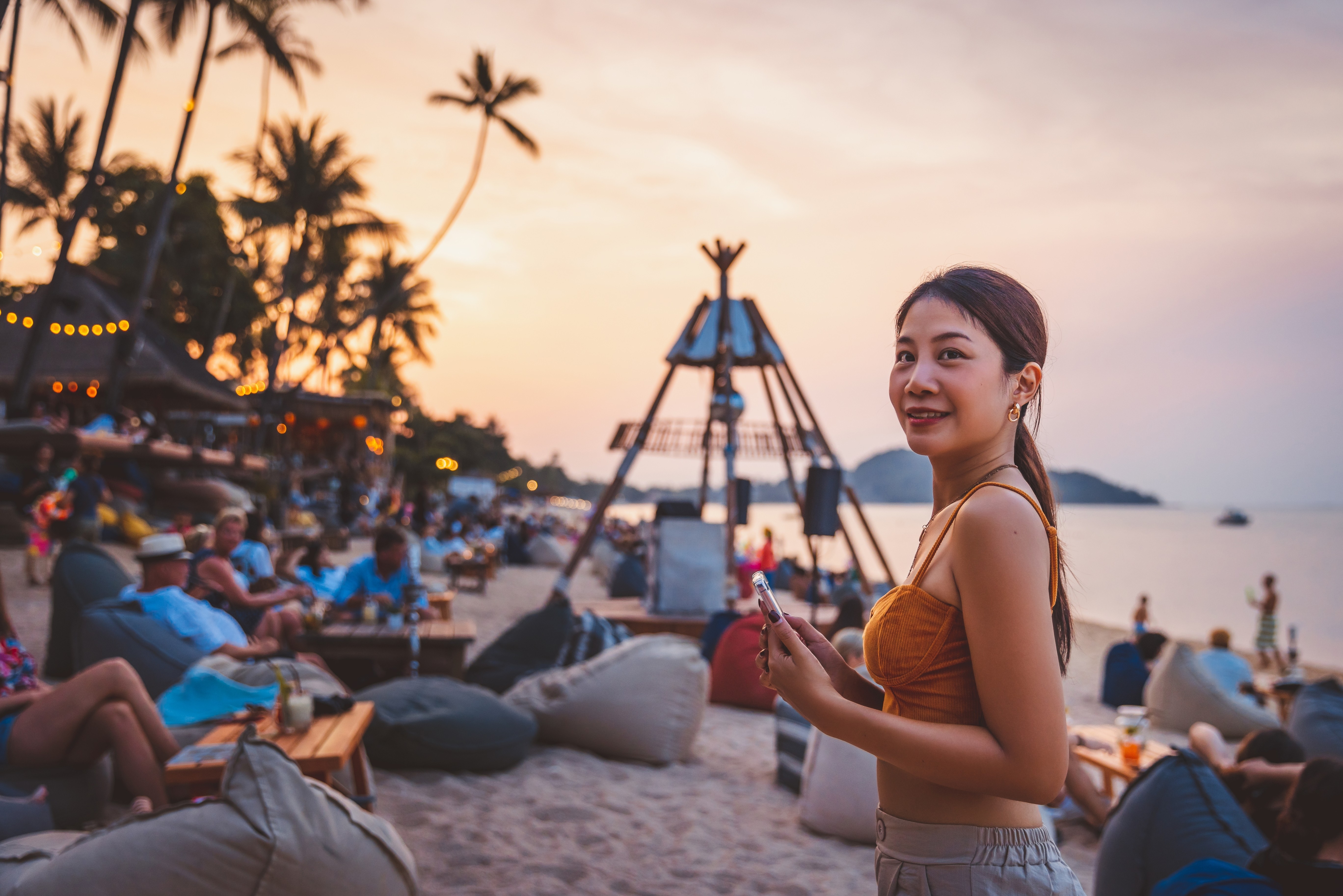 A woman on a busy beach at sunset, she smiles at the camera with her mobile phone in her hand
