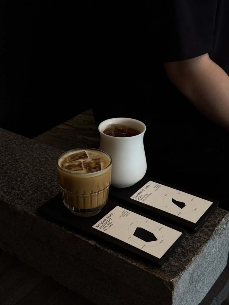 A glass of iced coffee alongside a white porcelain cup of tea, placed on a stone surface with two menus featuring geometric designs.