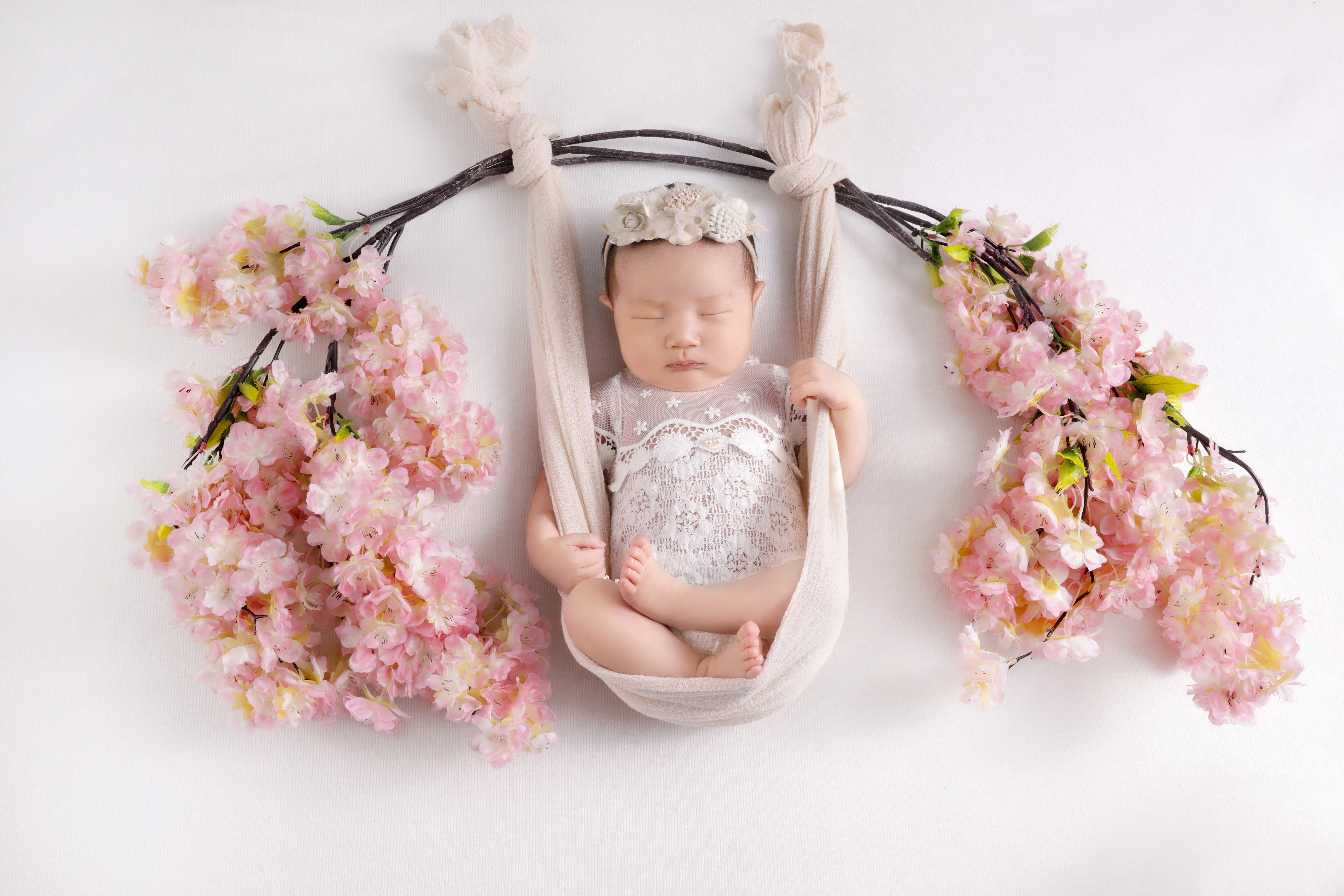 An infant photoshoot featuring a newborn baby peacefully sleeping in a delicate hammock adorned with soft pink cherry blossoms.