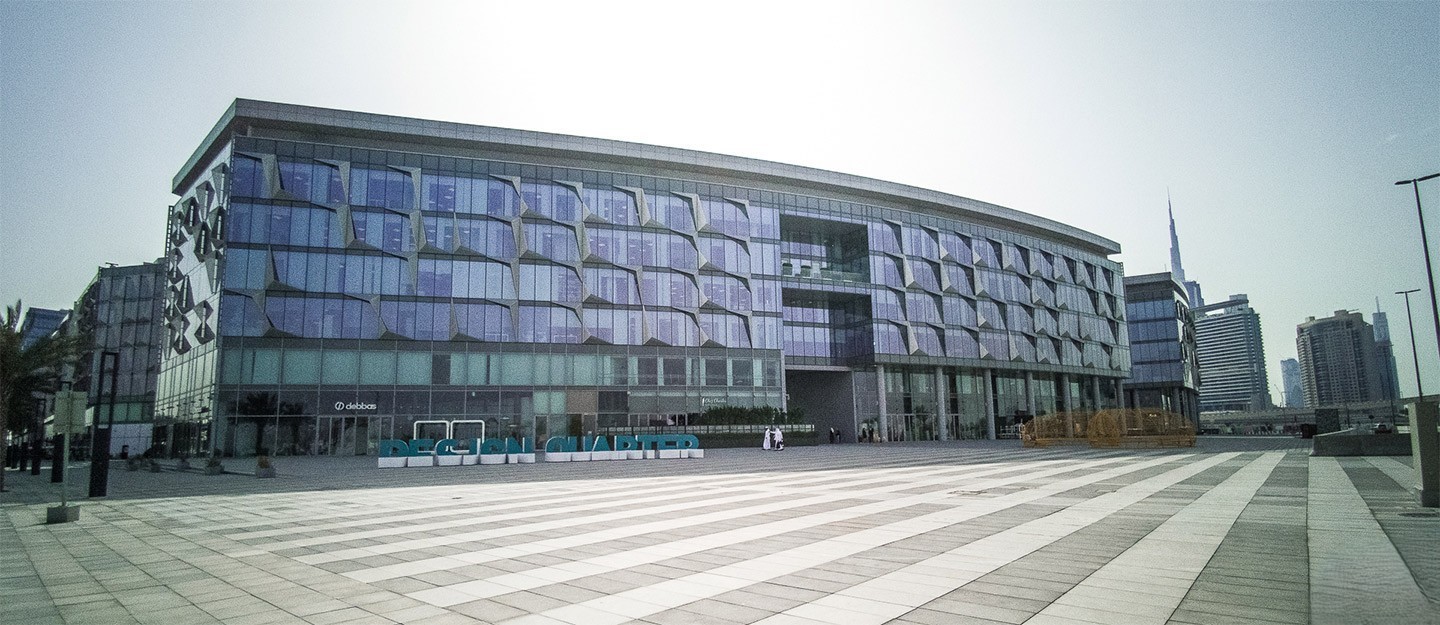 The modern, blue-glass-fronted Dubai Design District (d3) building with distinctive window patterns and a large paved plaza foreground.