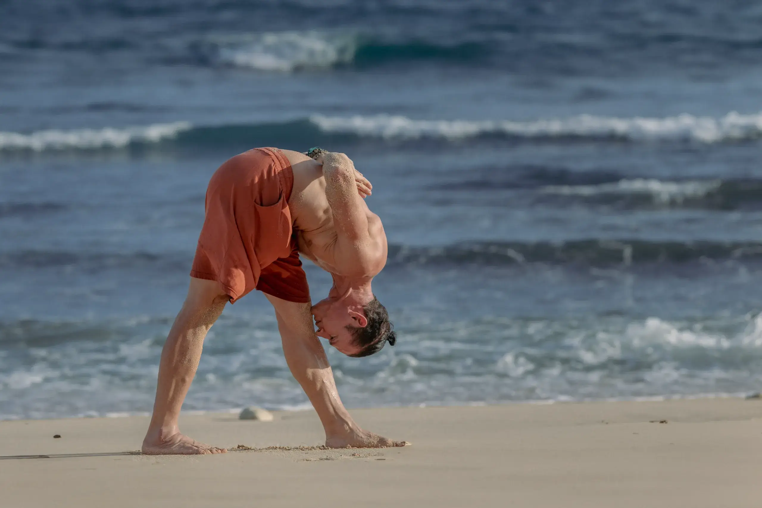 Advanced practitioner performing wide-legged forward fold with shoulder stretch on the beach for 300-hour yoga certification