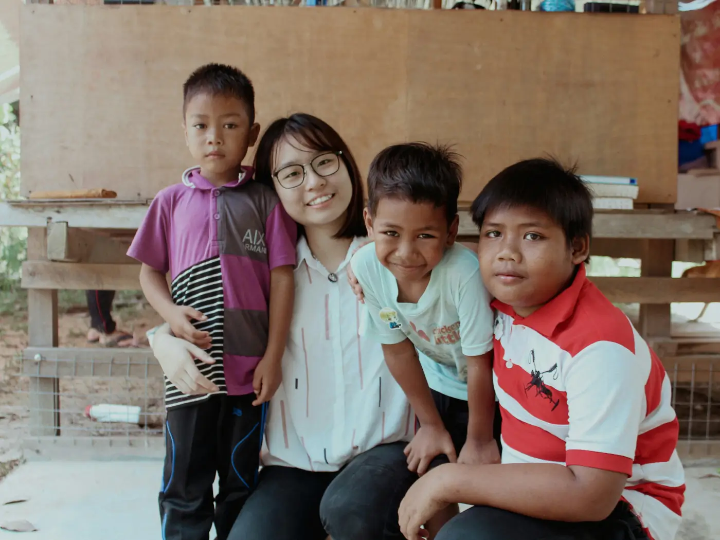 A group of three children and a young girl smiles together indoors, sitting on a bench in a casual setting.