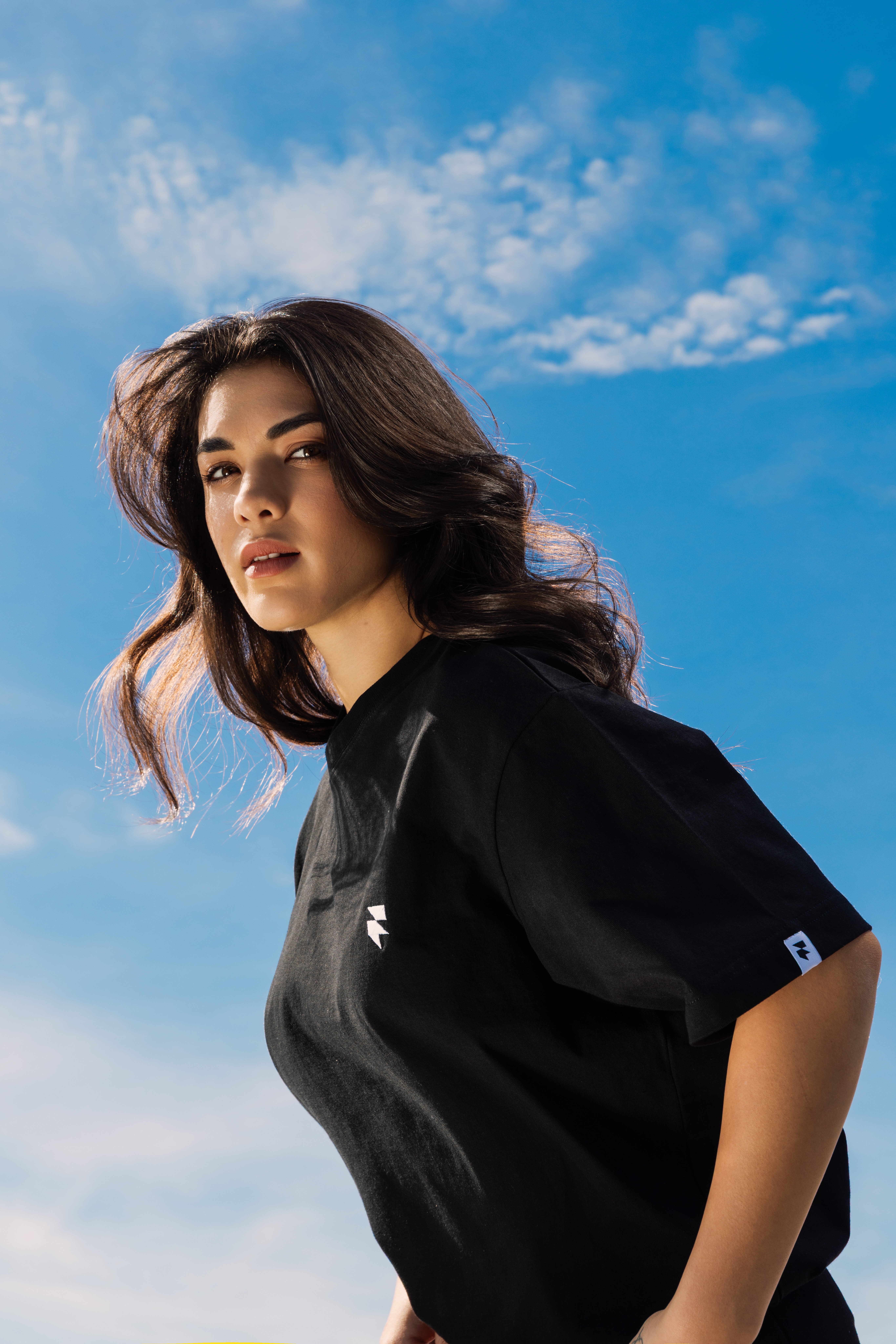 A woman with long, wavy hair wearing a black T-shirt poses confidently against a vibrant blue sky with scattered clouds.